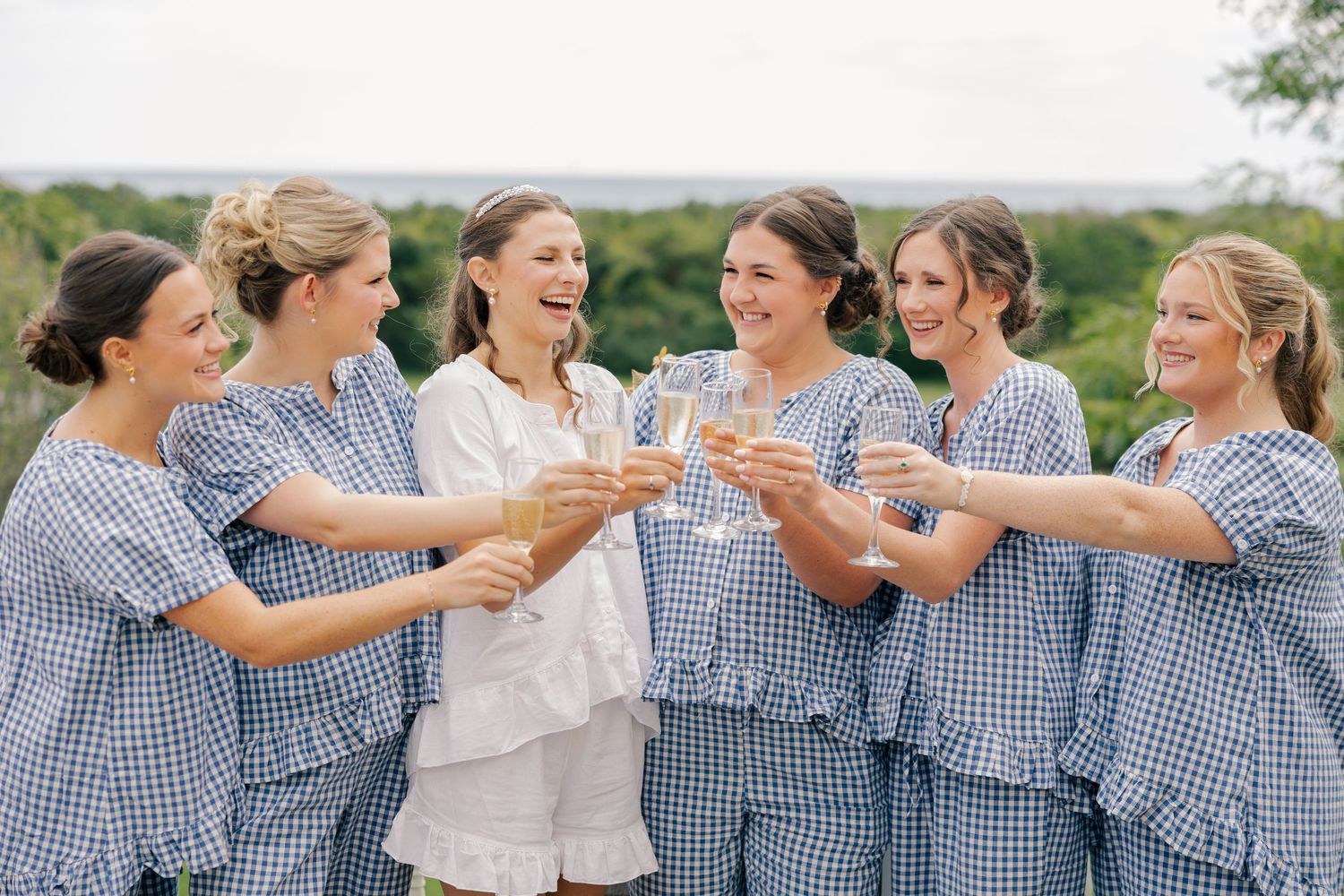 A bride in white pajamas toasts champagne with six bridesmaids wearing matching blue gingham sets before the wedding.