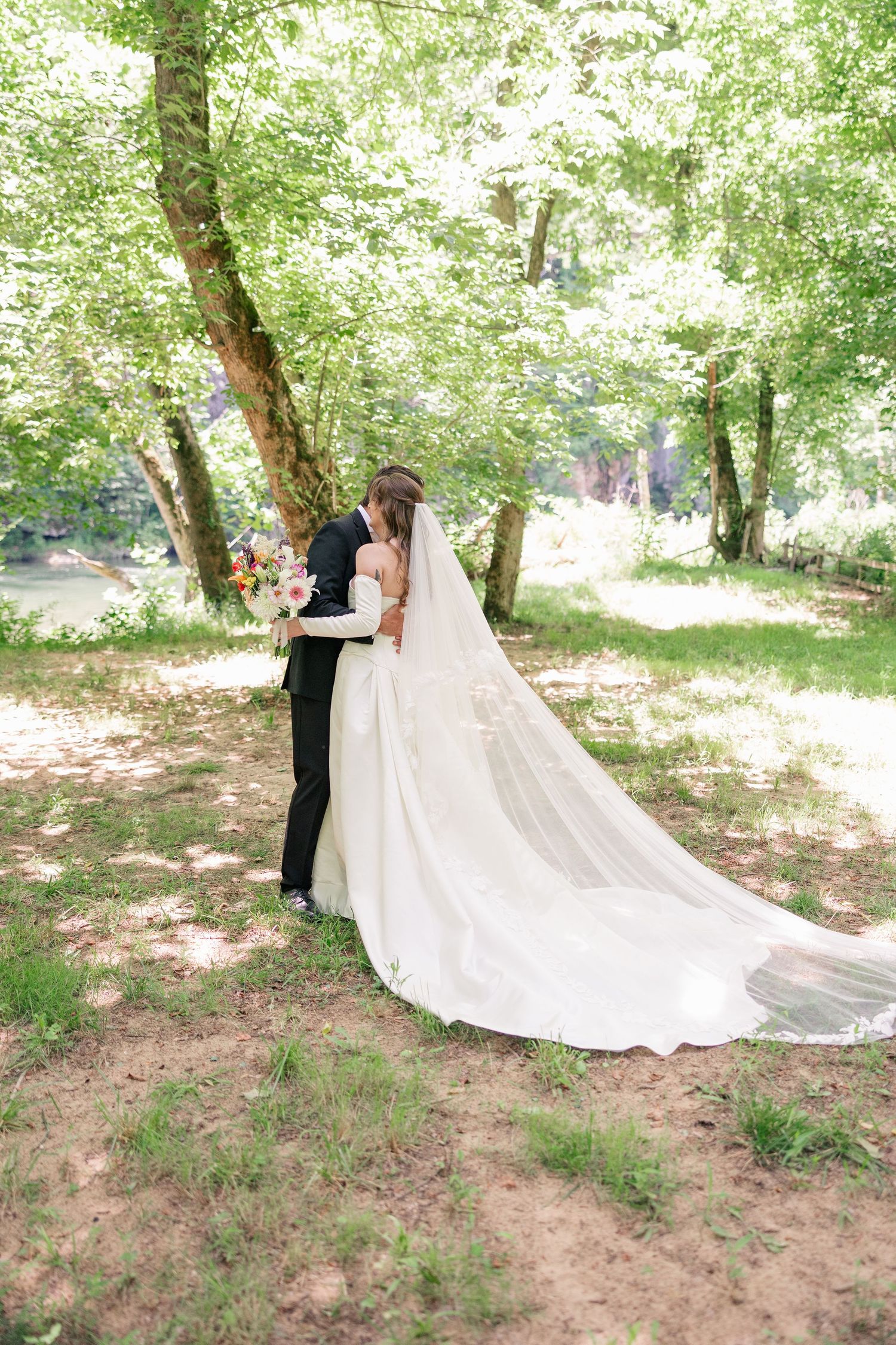 Bride and groom share a tender embrace under lush green trees, her cathedral veil flowing gracefully.