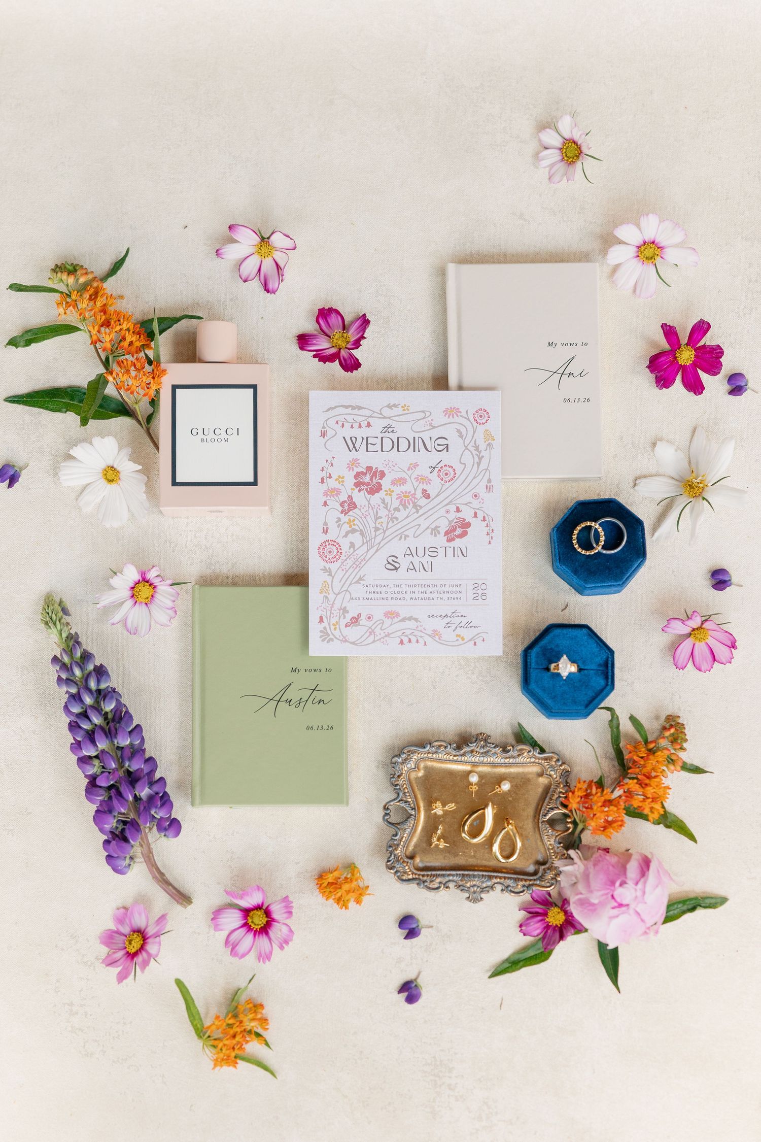 Elegant wedding flat lay with floral invitation, Gucci perfume, gold rings in blue velvet box, earrings, and colorful wildflowers.