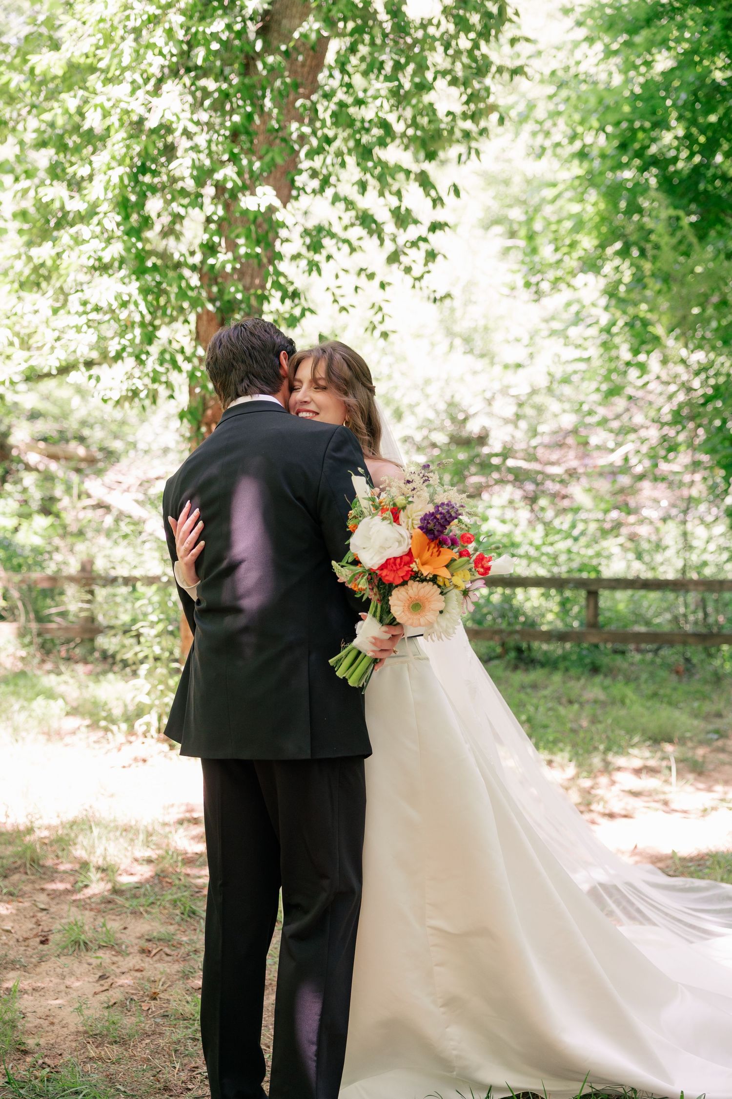 Bride and groom share an intimate embrace under a lush green tree, bride holding a vibrant colorful bouquet.