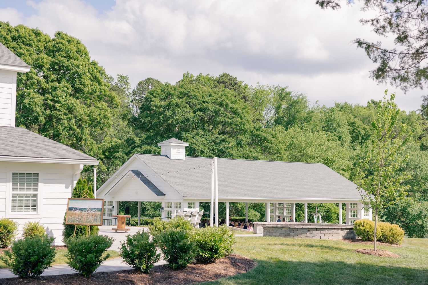 White country estate with covered pavilion surrounded by lush green trees and manicured shrubs on a sunny day.