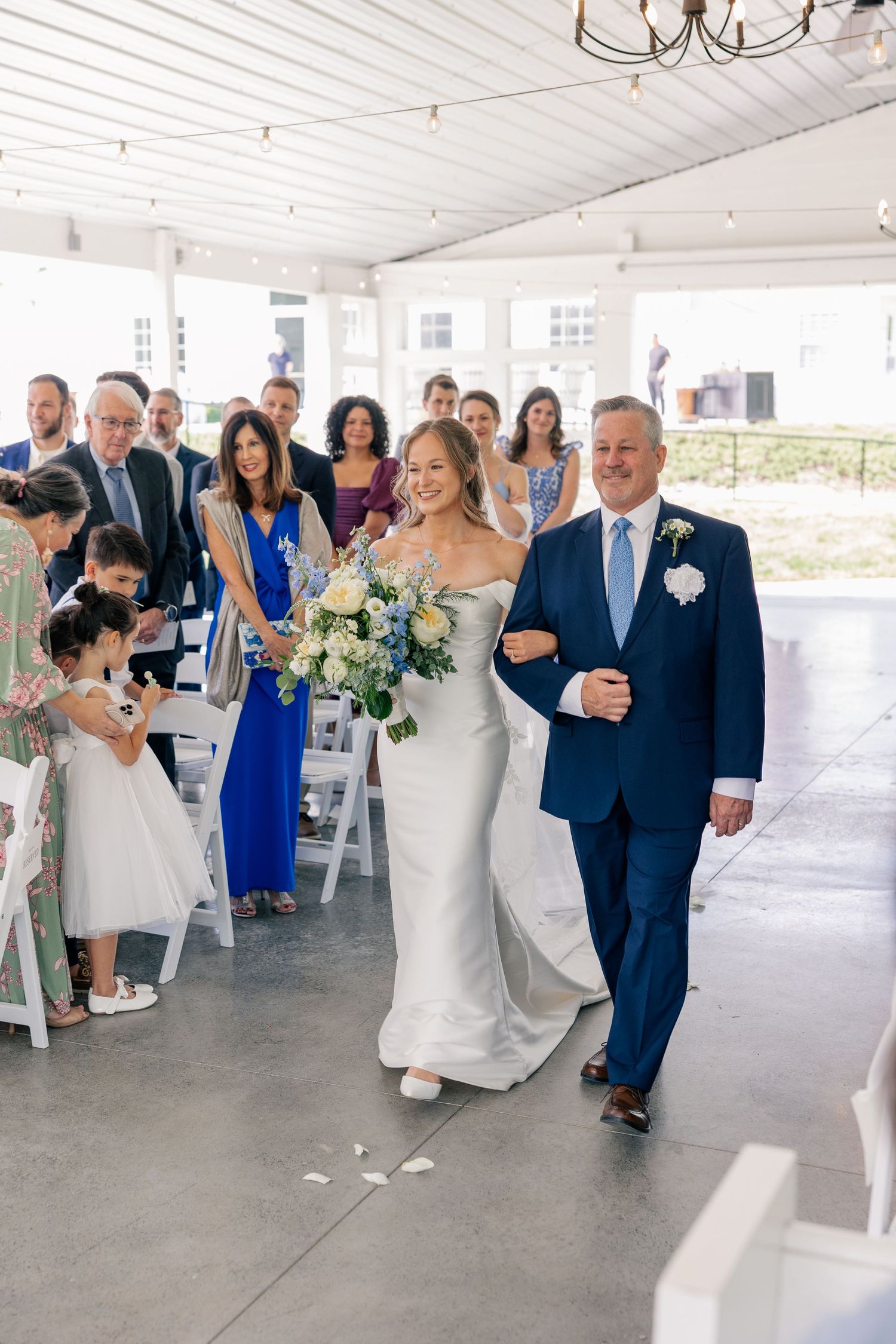 Bride walks down the aisle with her father in a white wedding gown holding a blue and white bouquet.