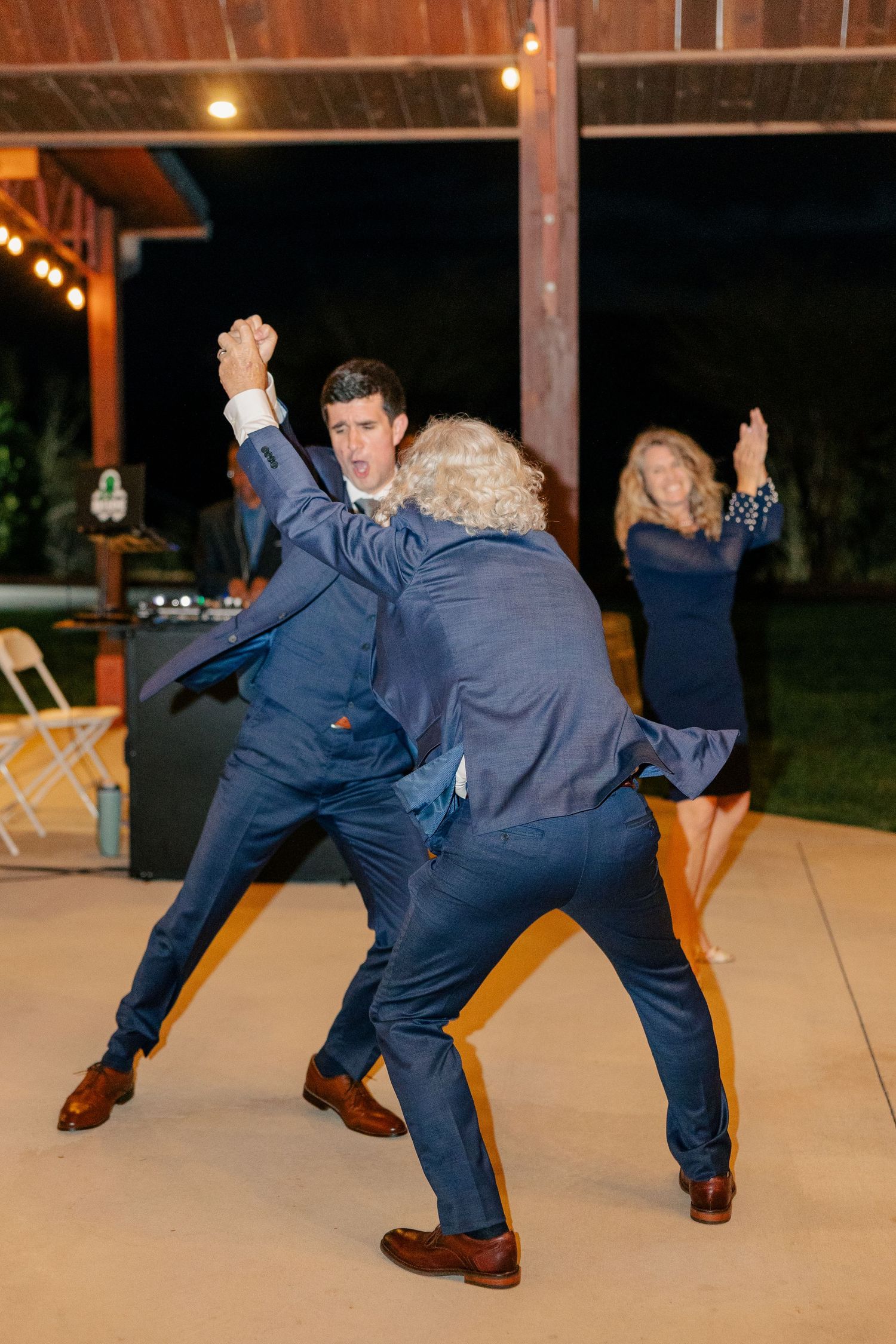 Groomsmen in navy suits dancing energetically at an outdoor evening wedding reception celebration.