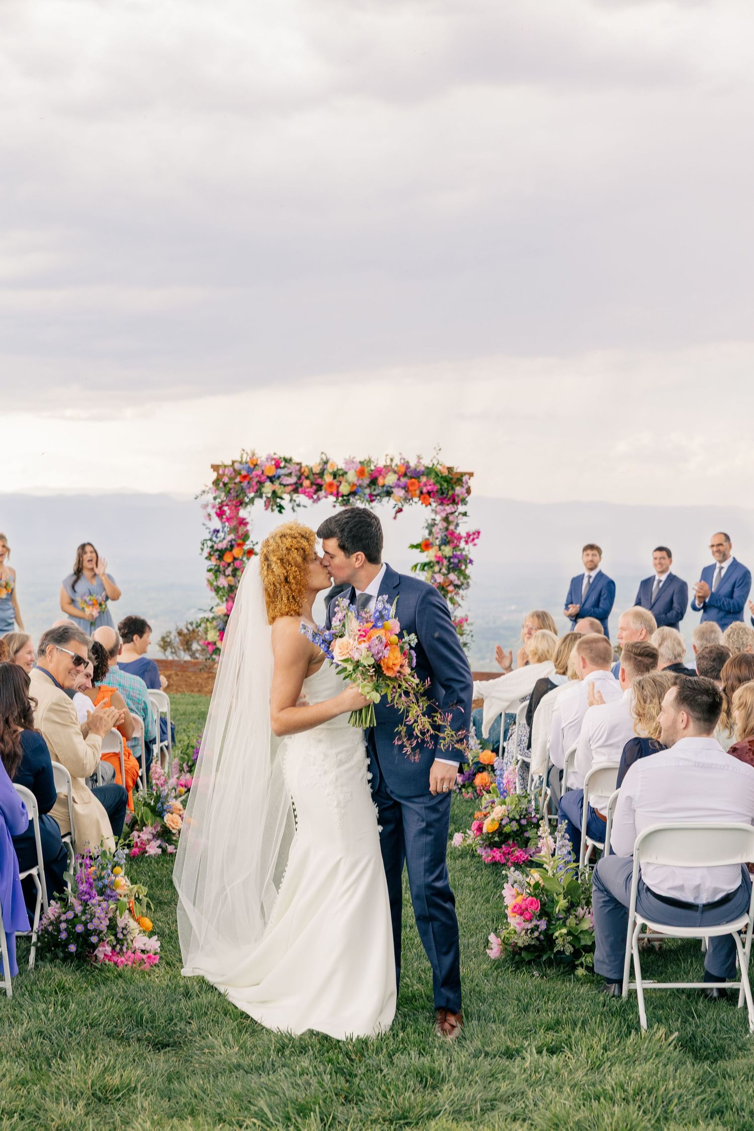 Bride and groom share first kiss at outdoor ceremony with colorful floral arch and seated wedding guests.