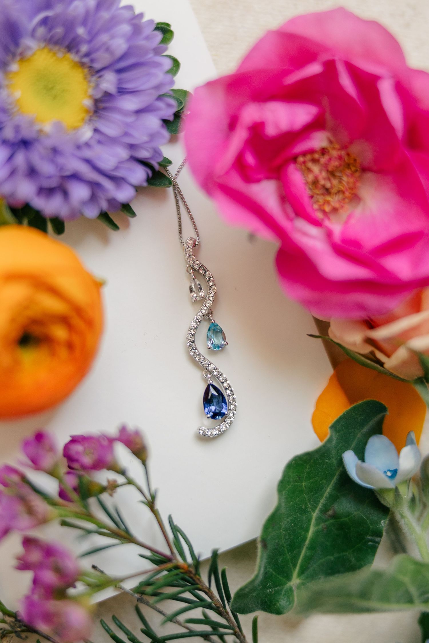 Elegant diamond and sapphire pendant necklace surrounded by colorful pink, purple, and orange flowers.