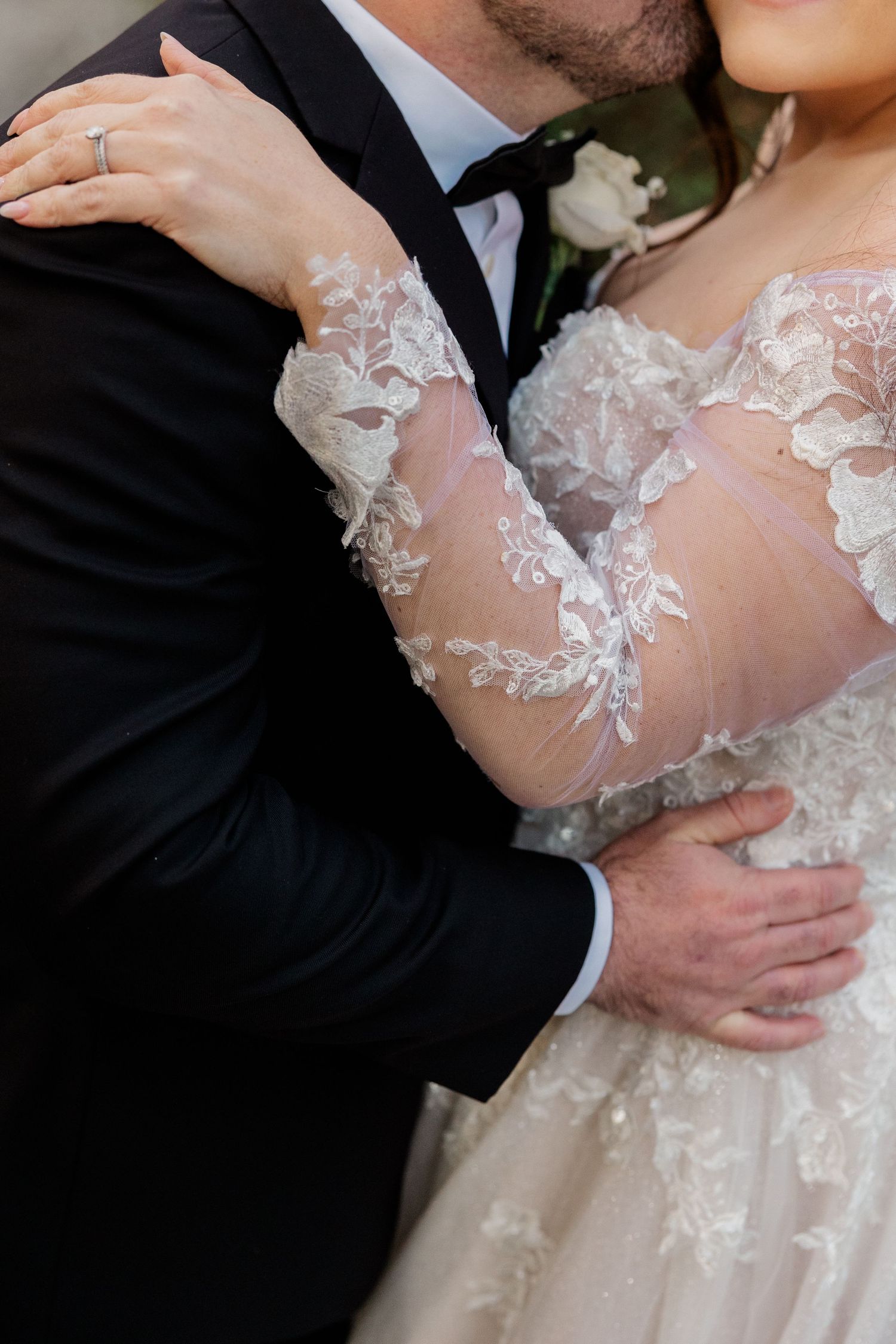 Close-up of newlywed couple embracing, showing intricate lace details on long-sleeved wedding dress and groom's suit. Wedding Photographer