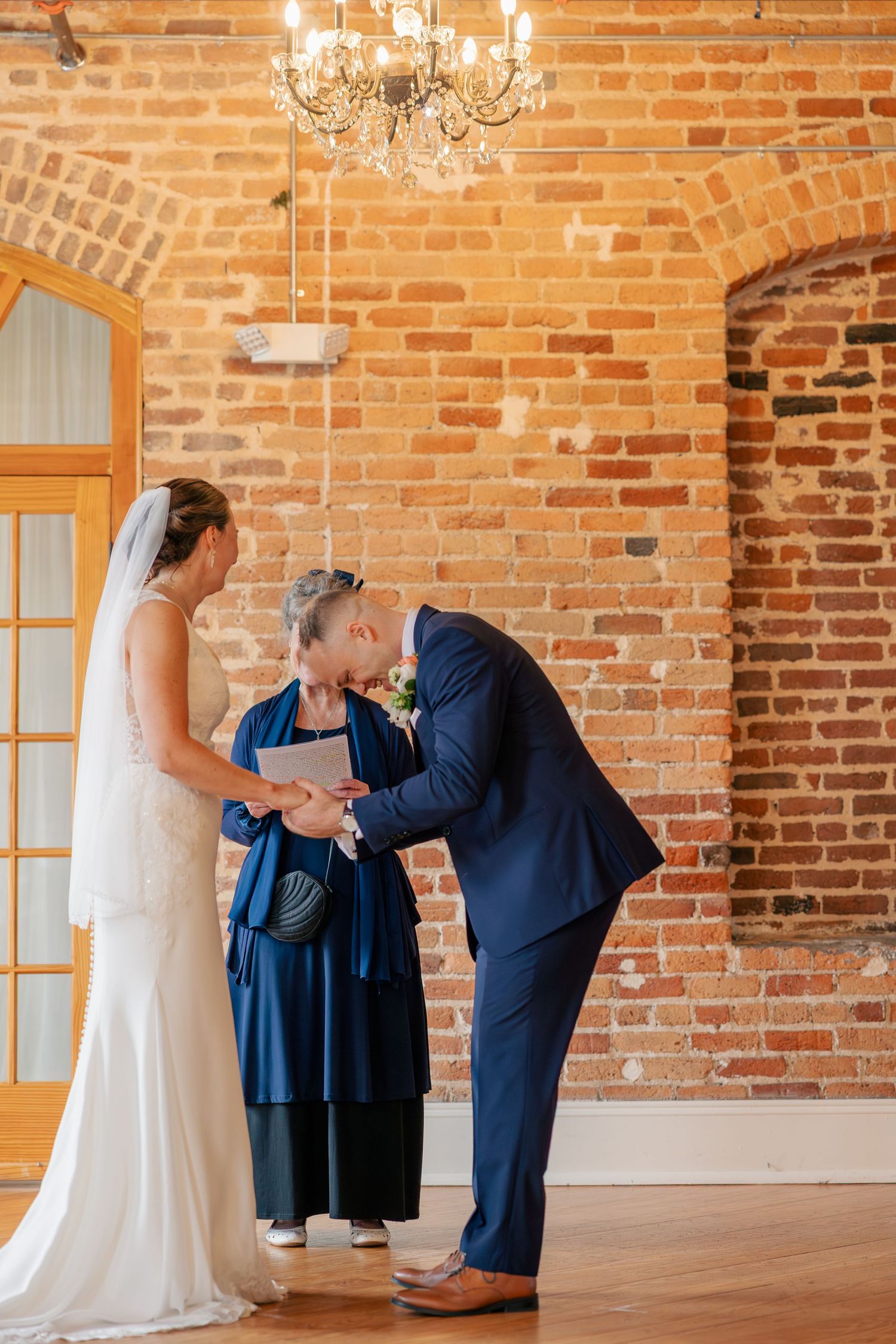 Bride and groom exchange vows with officiant during intimate wedding ceremony in rustic brick venue with chandelier.