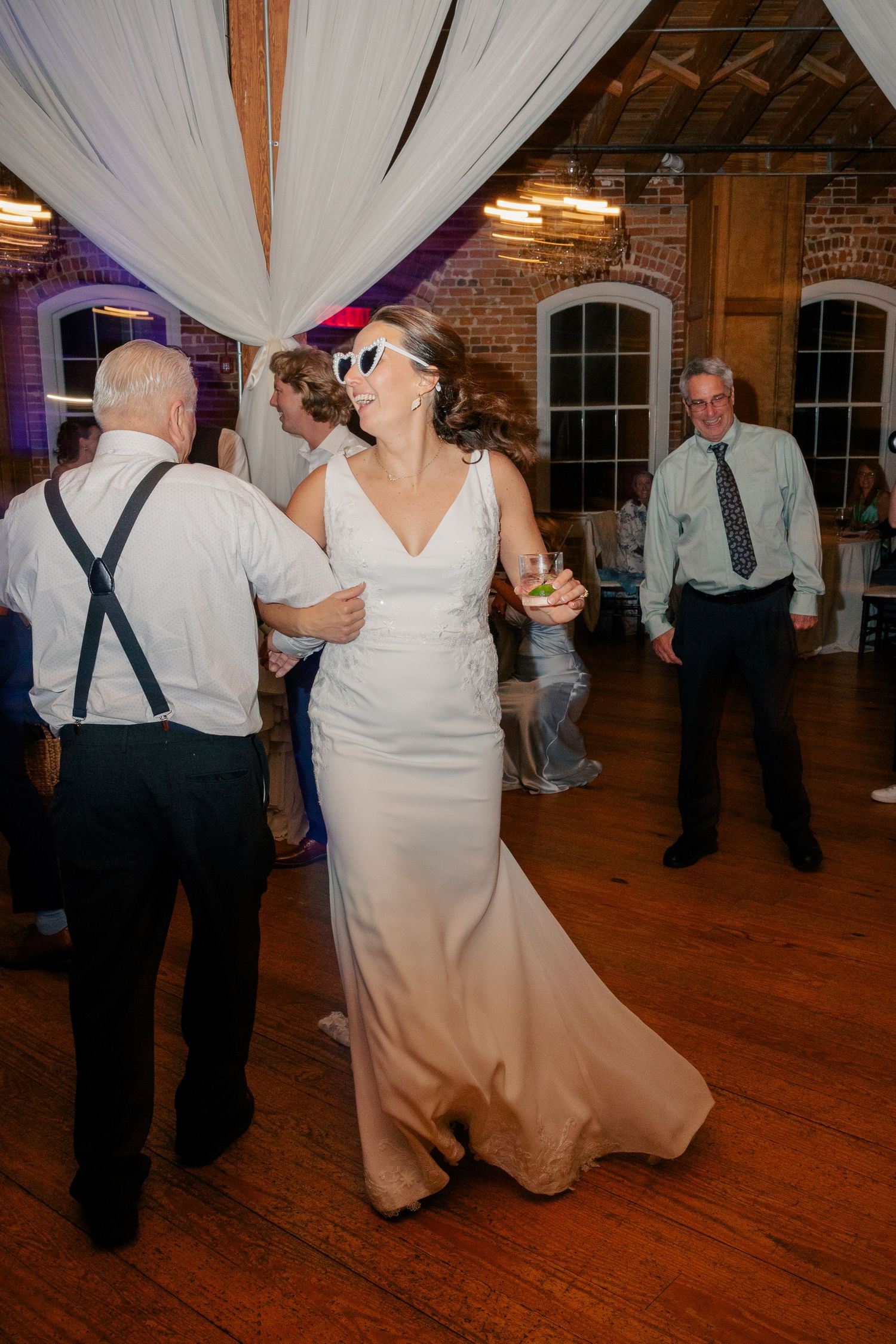 Bride in white gown wearing sunglasses dances joyfully with guests at wedding reception.
