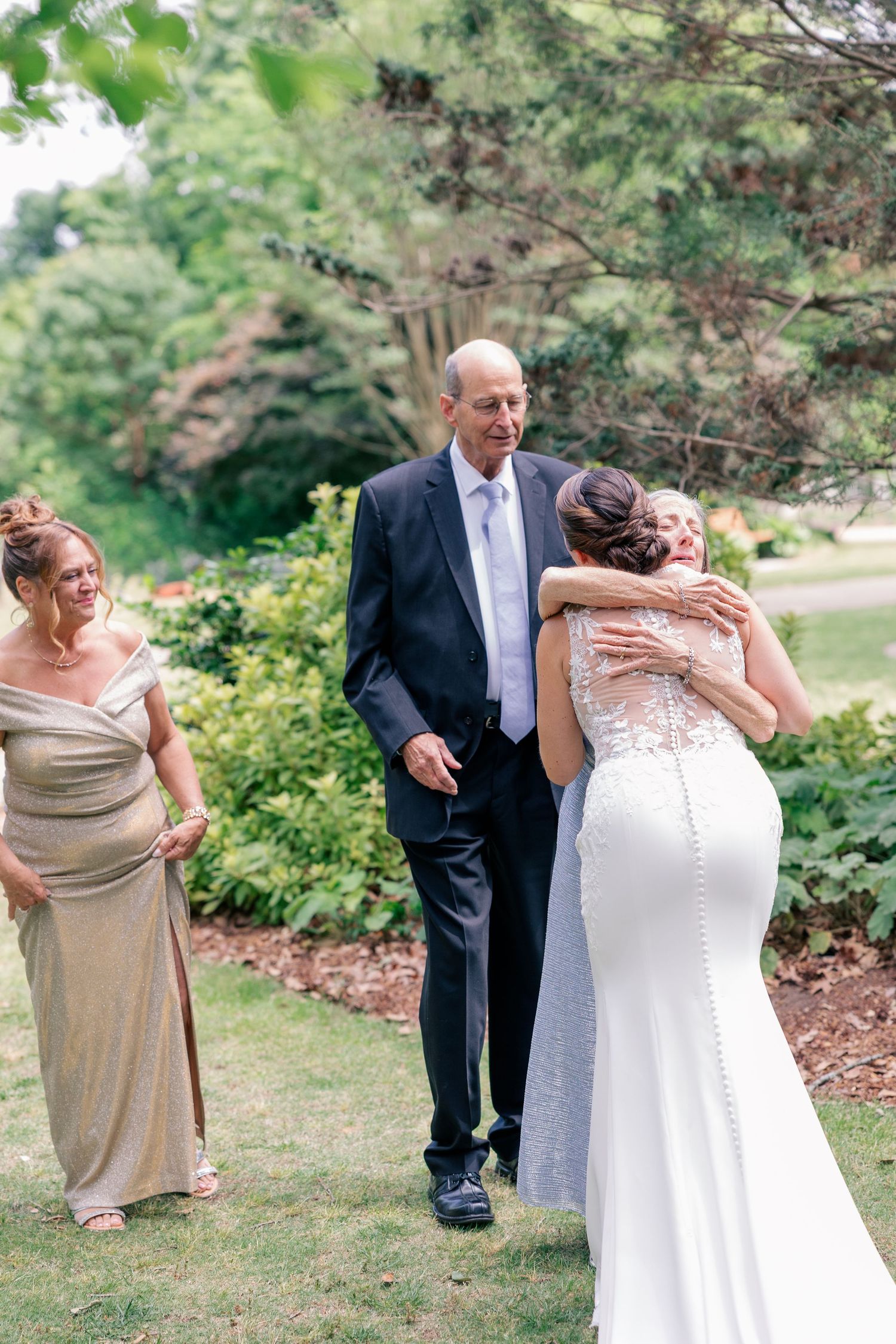 Bride embraces a family member while parents look on with joy in a beautiful garden setting.