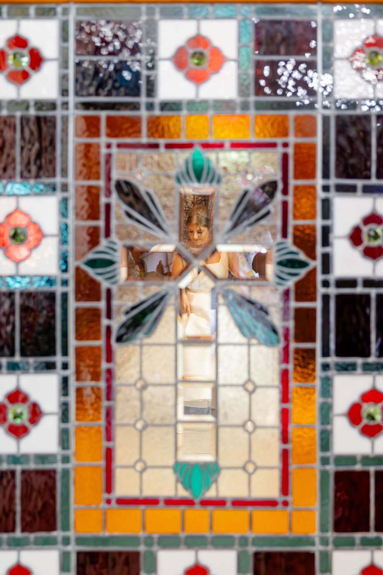 Bride reflected through colorful stained glass window featuring ornate cross in vibrant orange and teal hues.