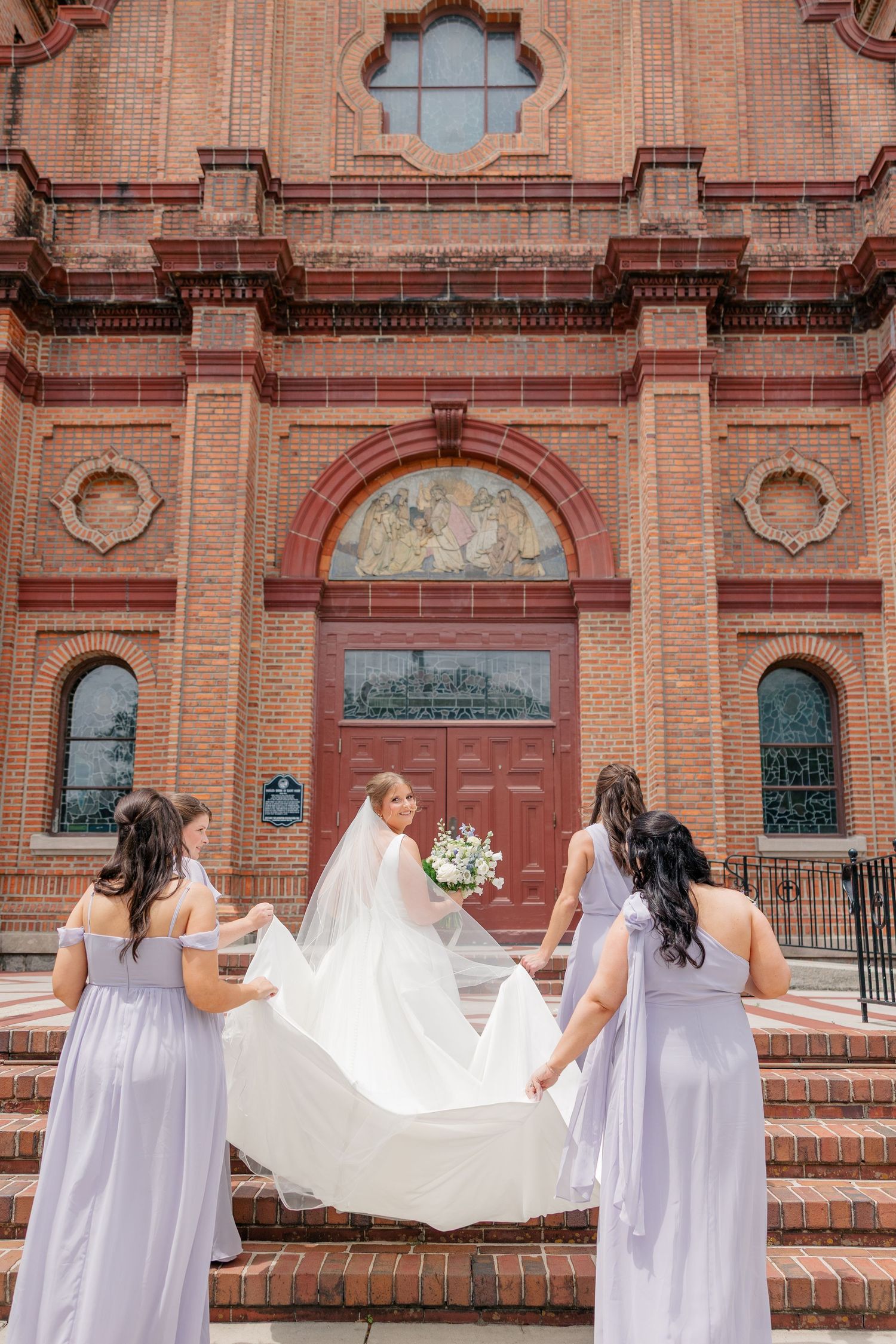 Bridesmaids in lavender dresses spread the bride's cathedral veil on the steps of a red brick church.