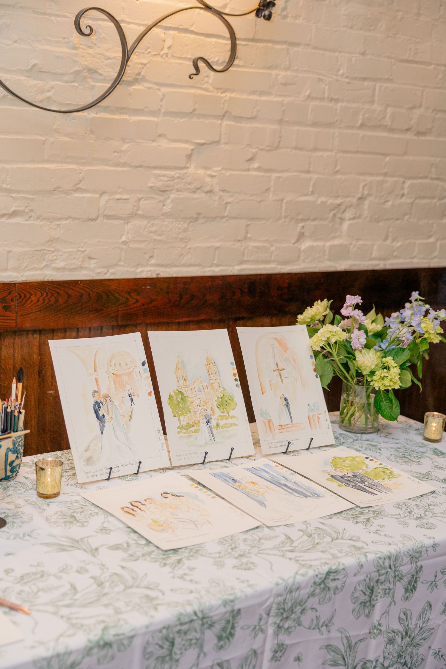 Live wedding painter displaying three watercolor illustrations of the couple and church on a toile-covered table.