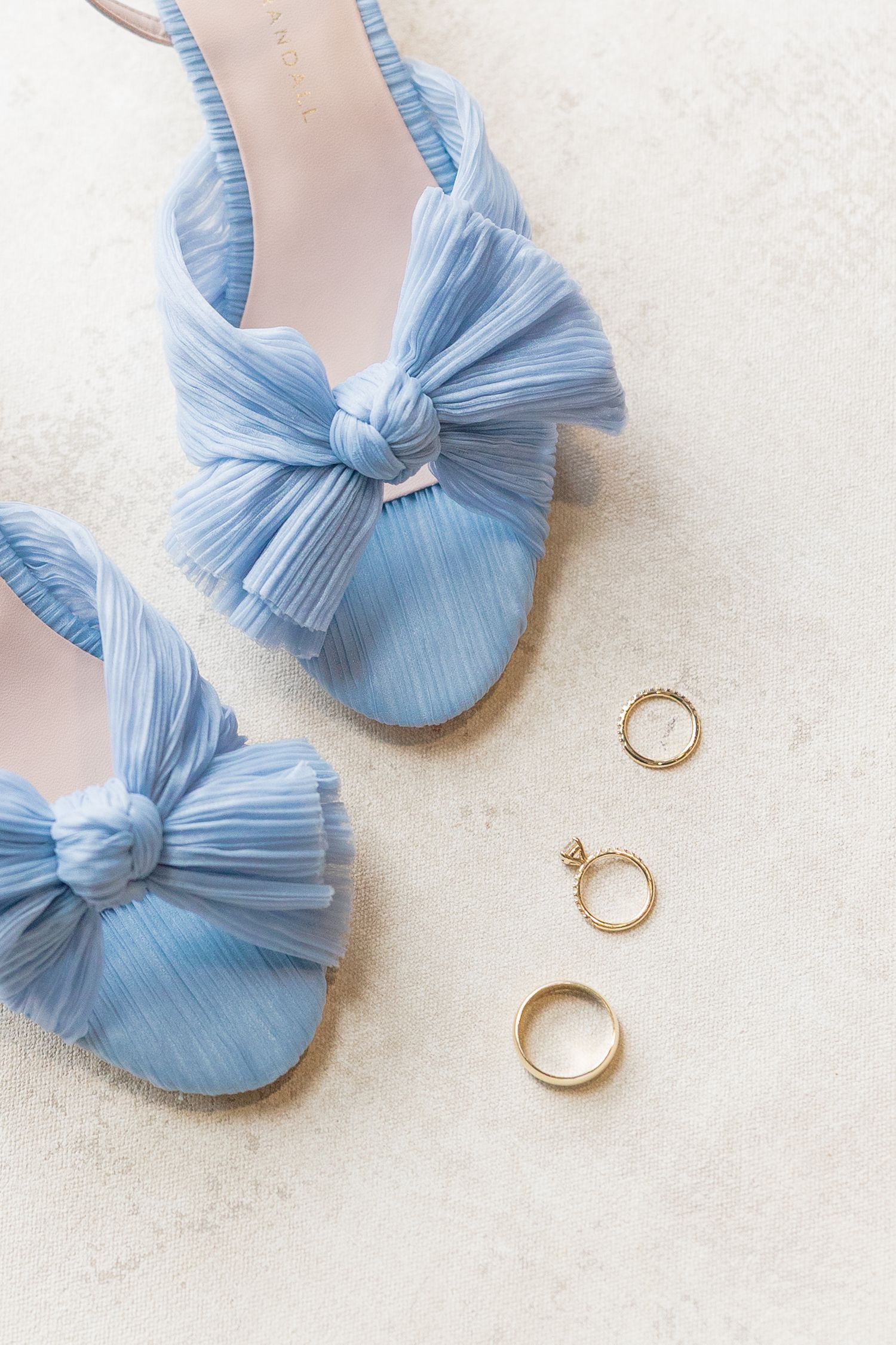 Light blue tulle bow heels displayed beside three gold wedding rings on a white marble surface.