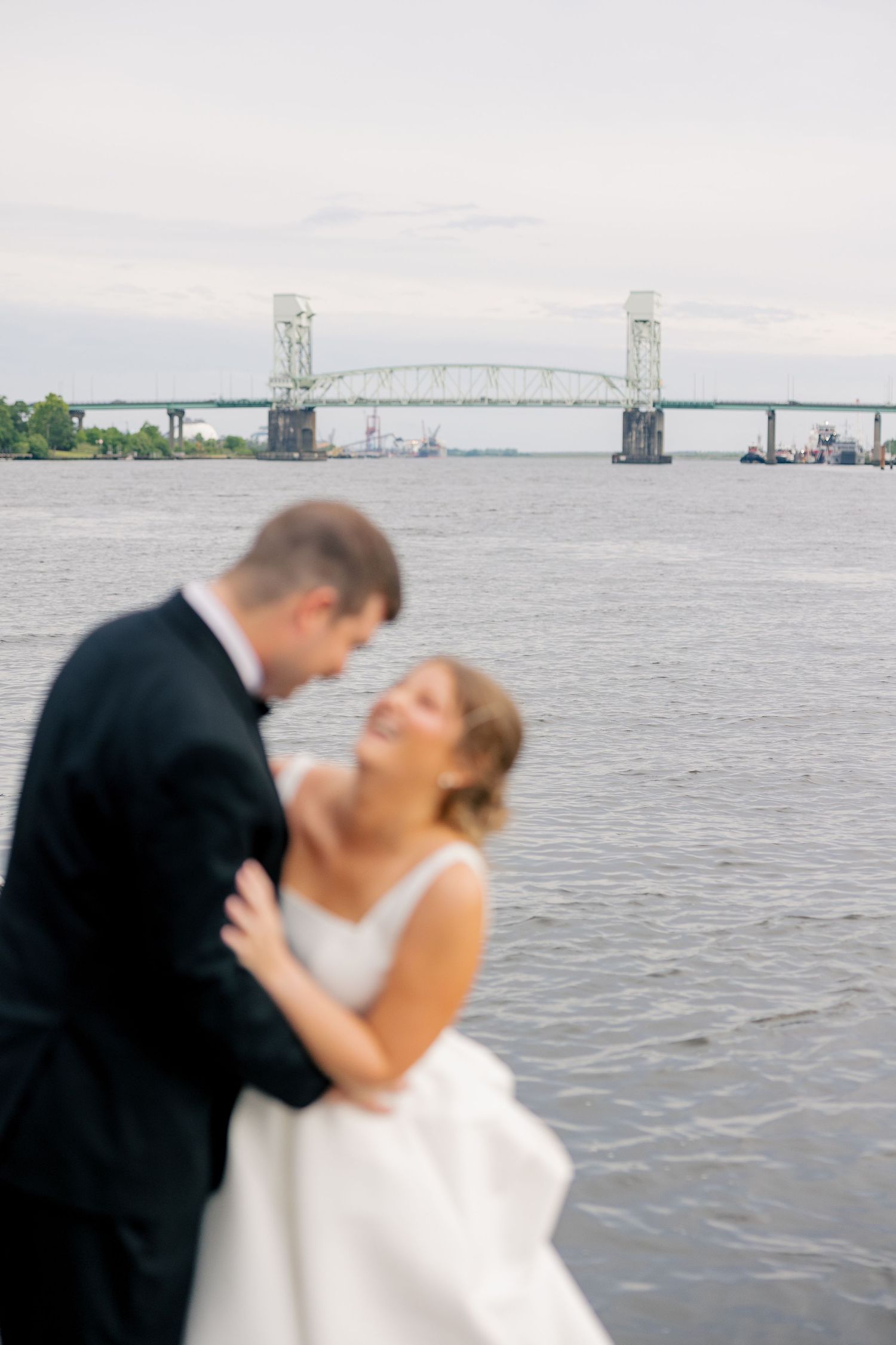 Blurred bride and groom embrace romantically with the Cape Fear Memorial Bridge over the river in the background.