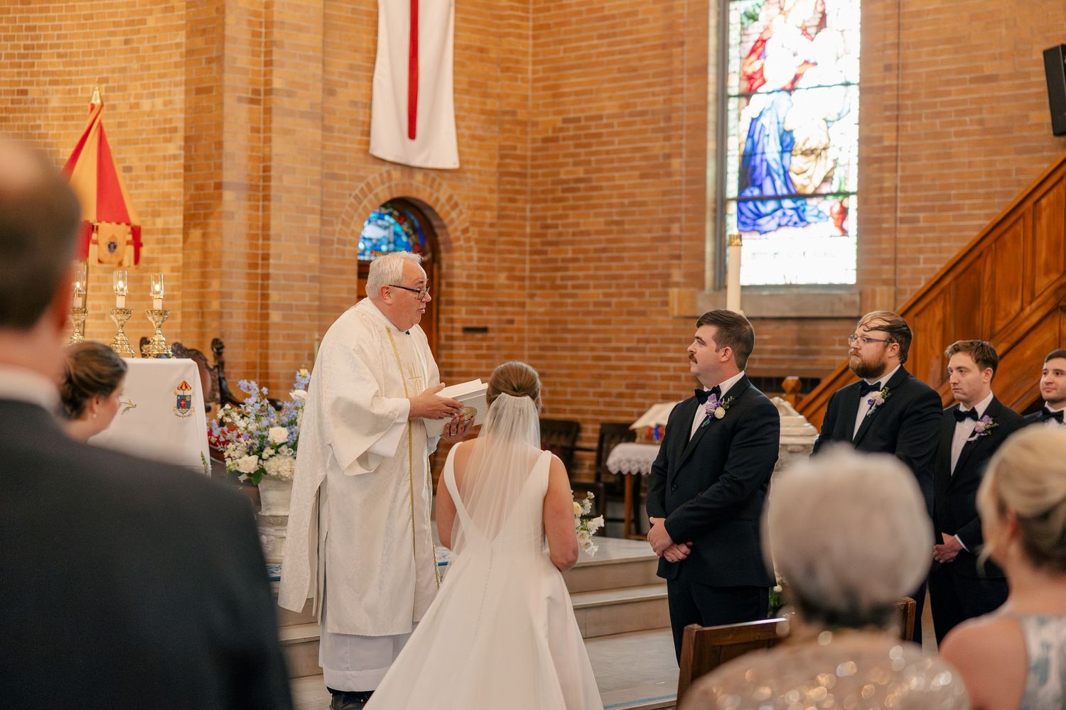 Priest officiates wedding ceremony at brick church altar with stained glass windows.