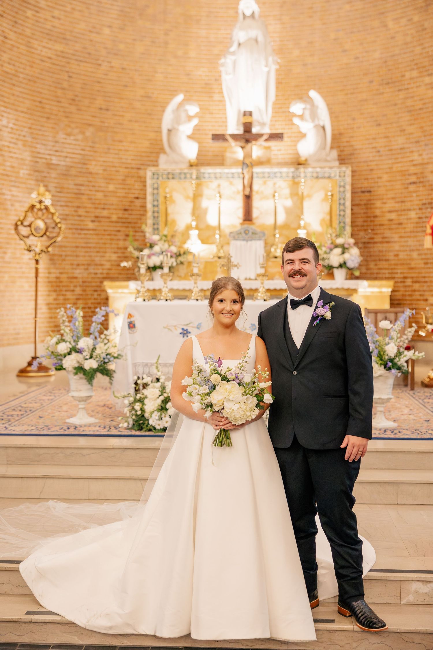 Bride and groom pose at ornate church altar, bride holding white floral bouquet in elegant gown.