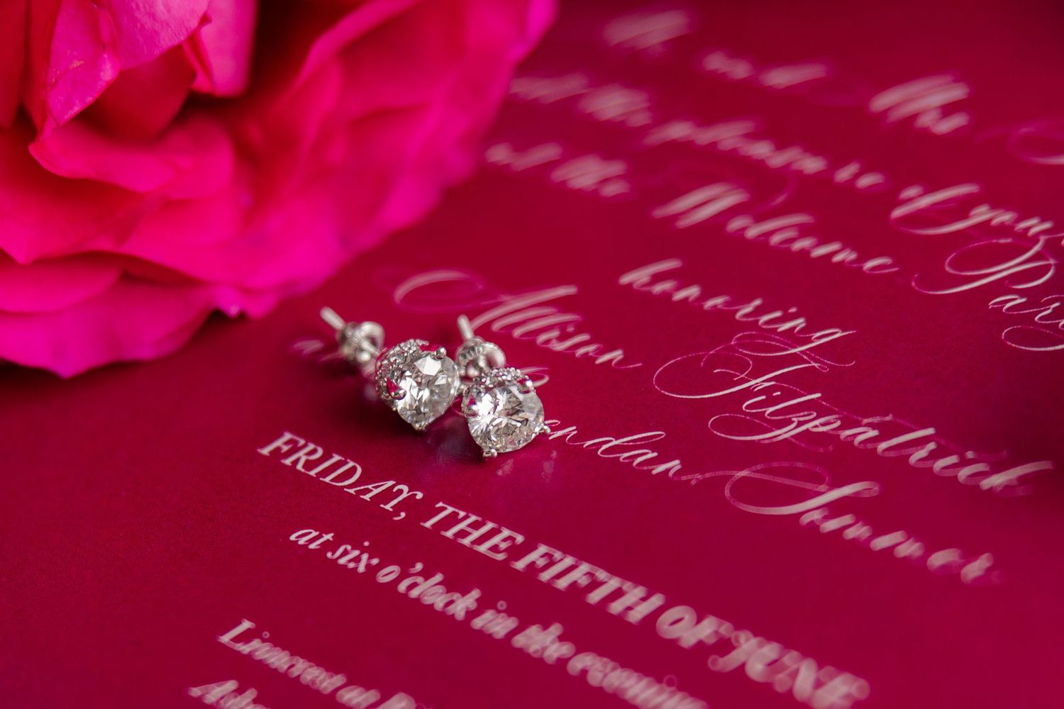 Diamond stud earrings on red wedding invitation with elegant white calligraphy script.