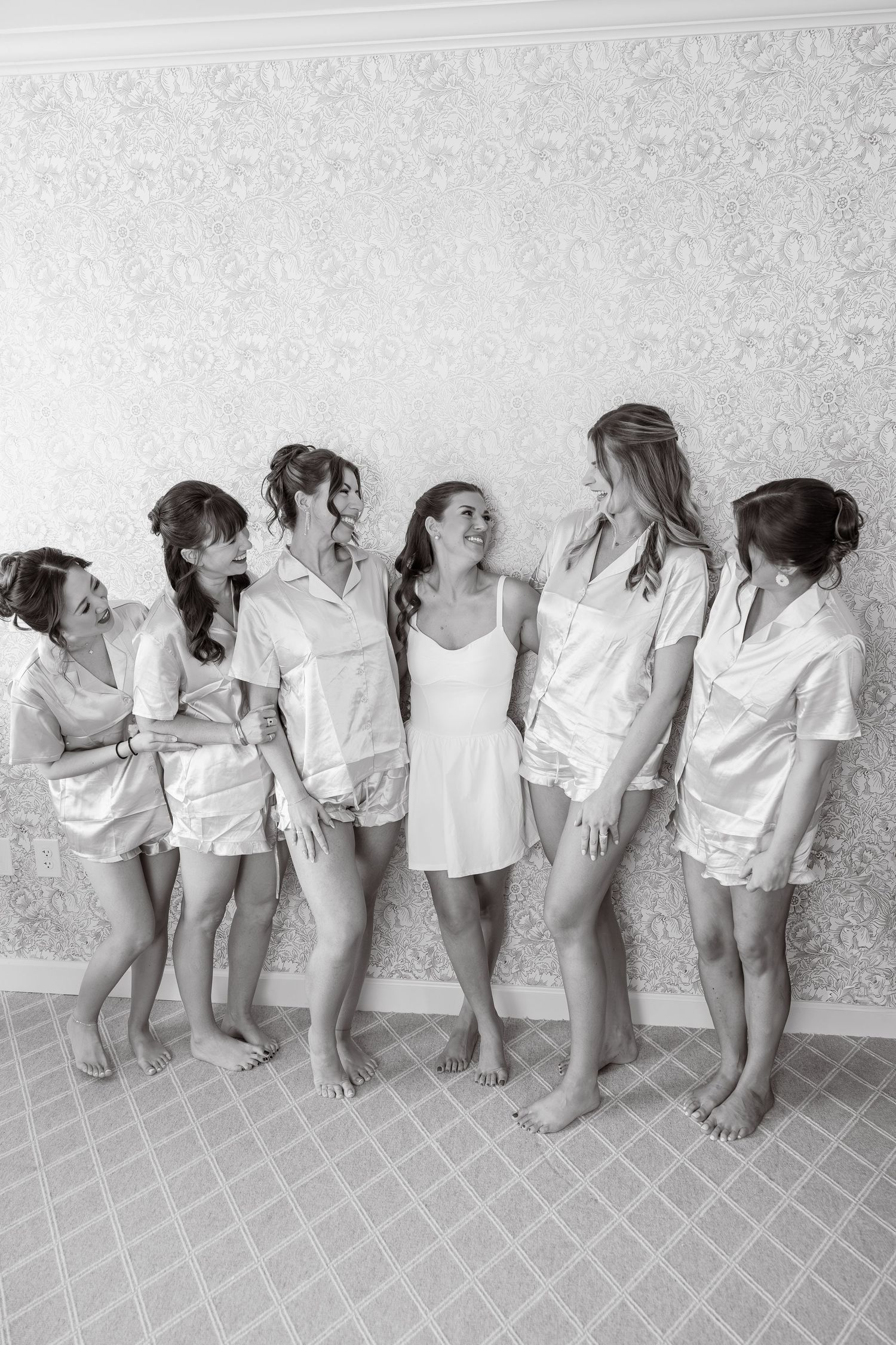 Bride and bridesmaids in matching satin pajamas laughing together during wedding morning preparations.