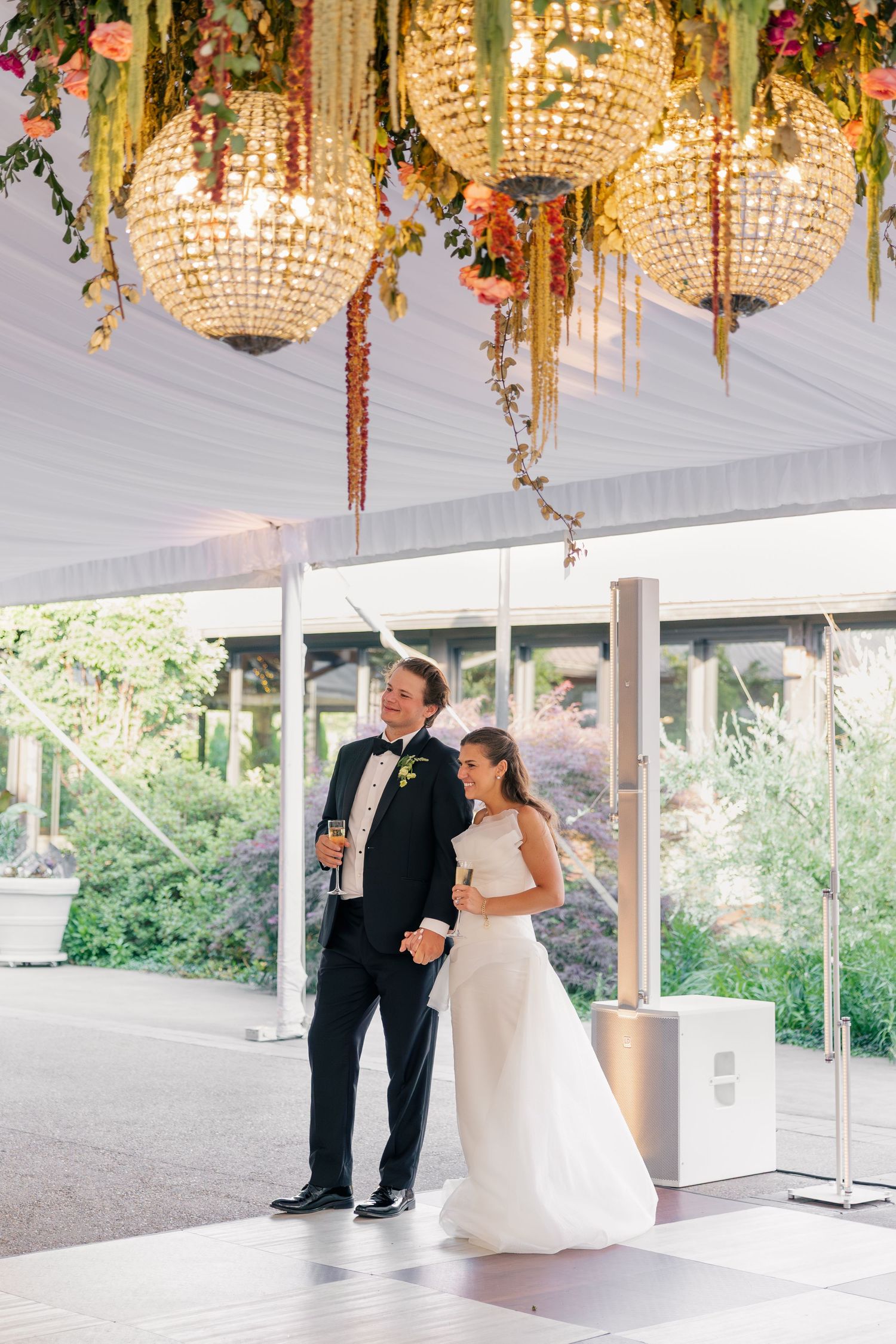 Bride and groom stand together under ornate chandelier in elegant outdoor tented reception.
