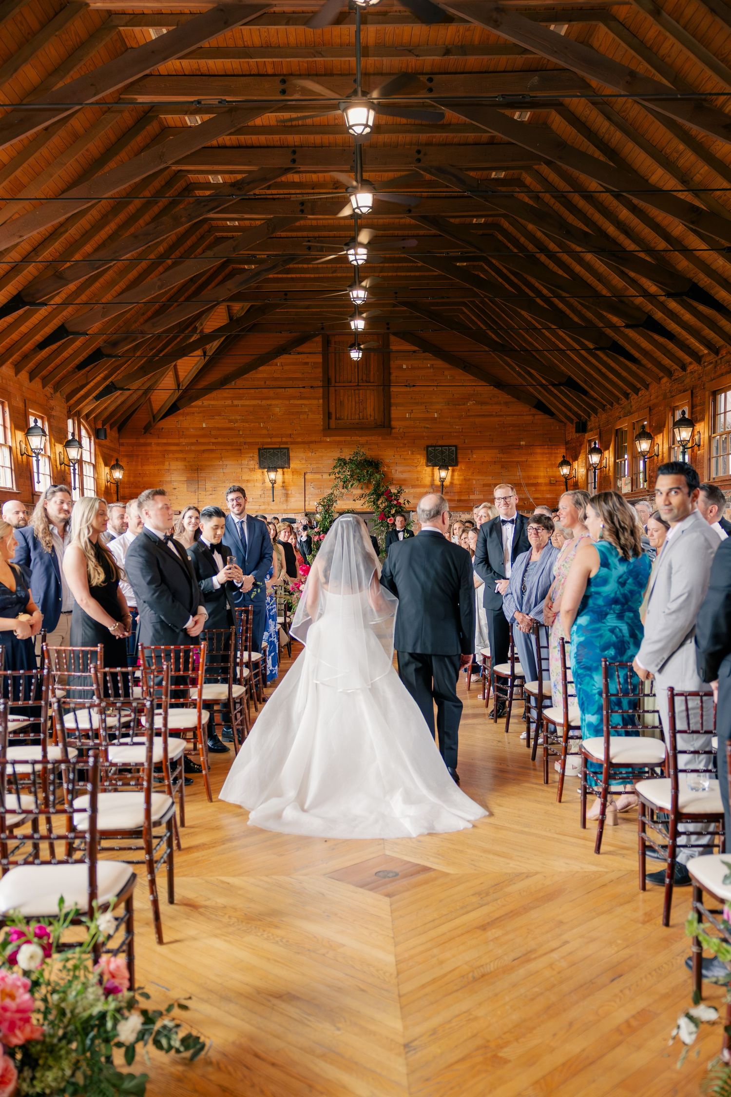 Bride walks down aisle in rustic wooden barn wedding venue with vaulted ceiling and seated guests.