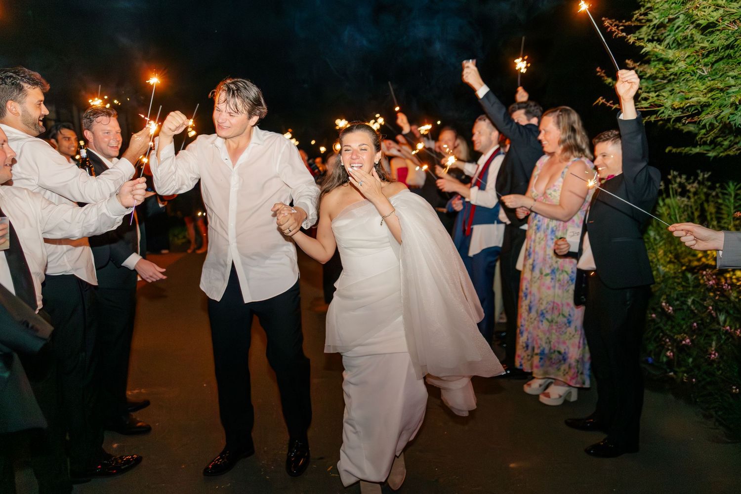 Bride and groom exit through a sparkler tunnel surrounded by cheering guests at night.