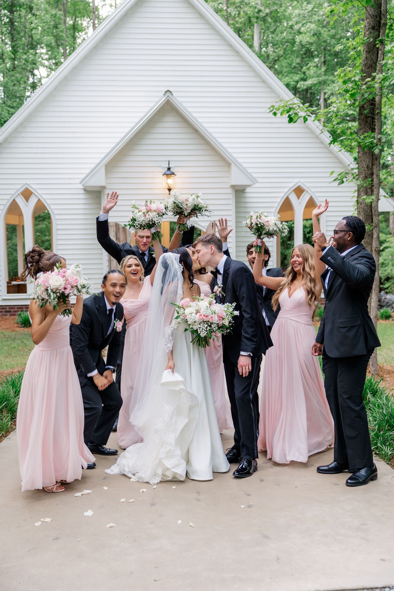 Wedding party celebrating outside a white chapel with bridesmaids in pink dresses.