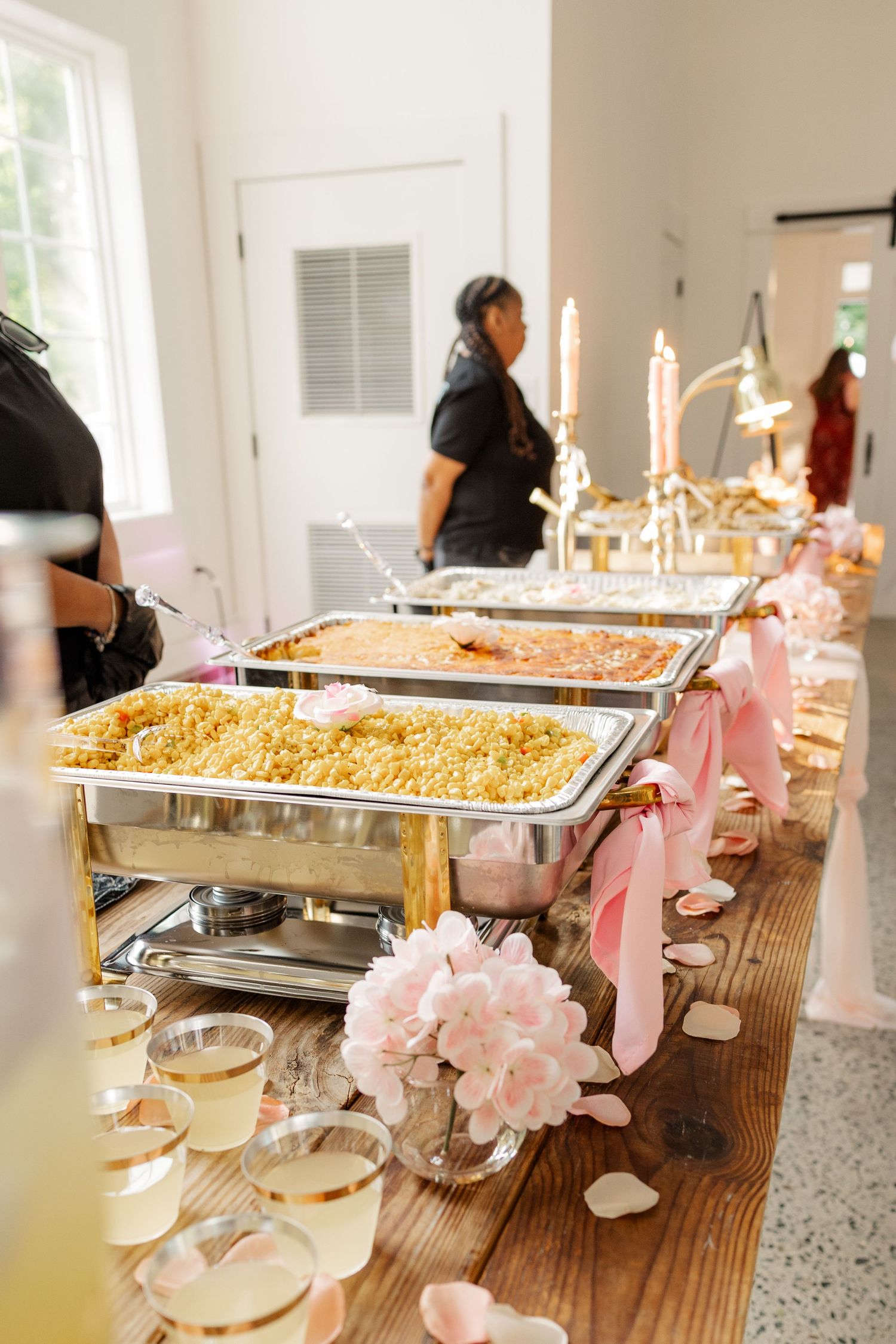 An elegant wedding buffet table decorated with pink floral accents and multiple chafing dishes of food.