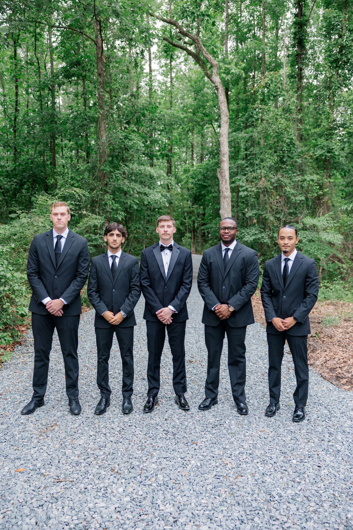 Groomsmen in black suits stand in a line on a gravel path surrounded by lush green trees.