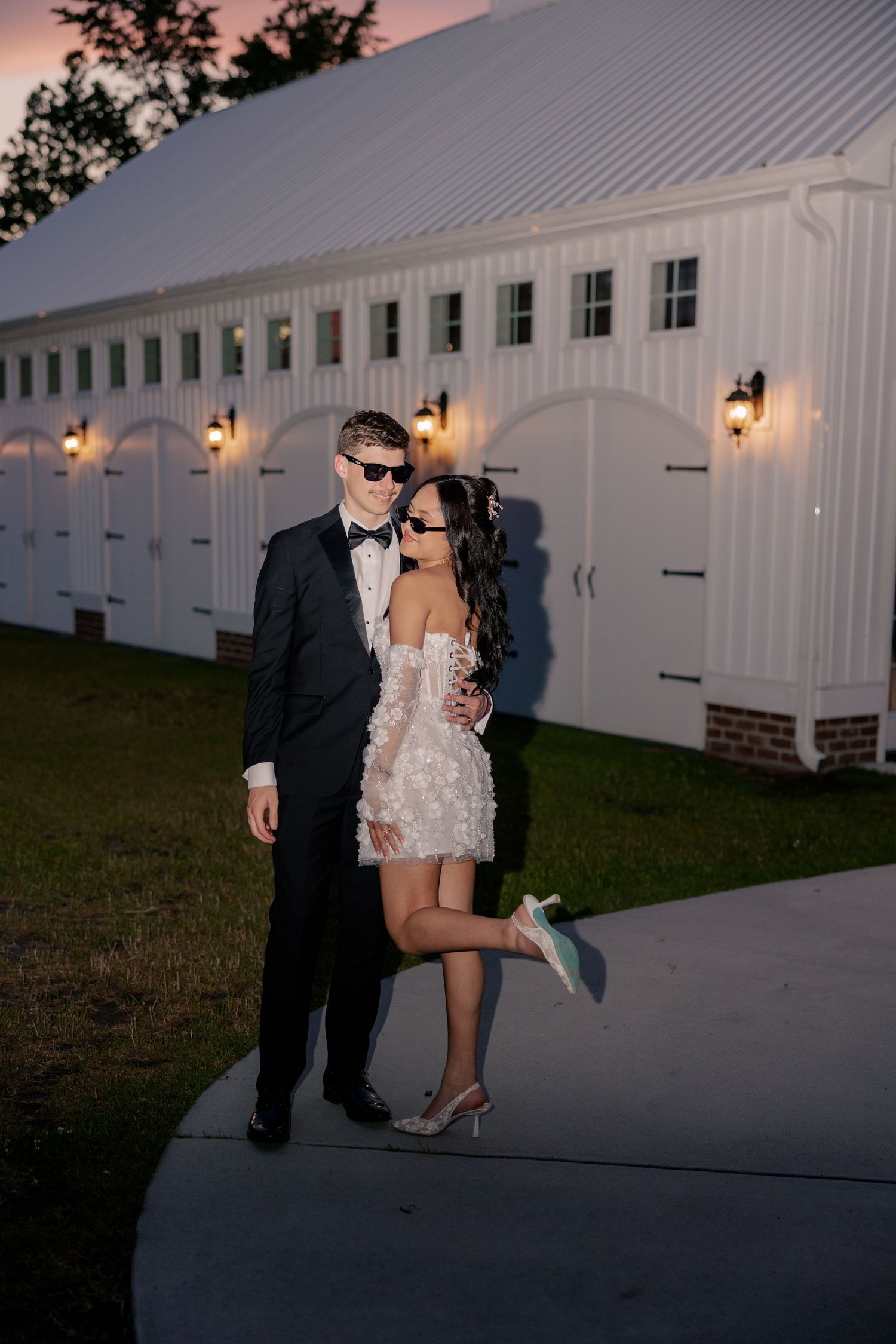 A couple shares an intimate moment outside a white barn-style building at sunset with warm lighting.