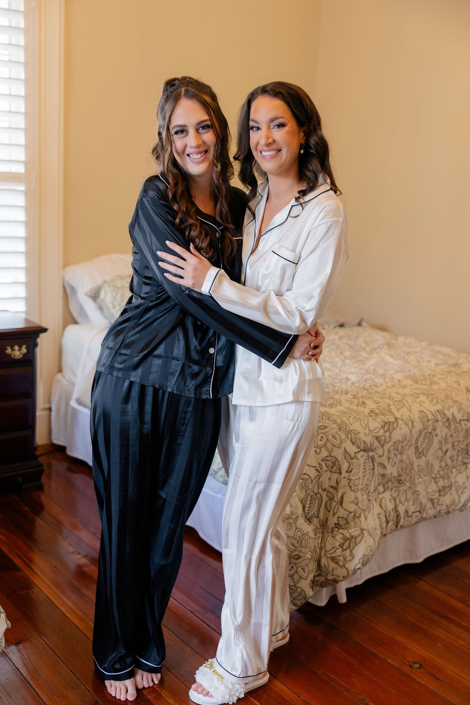 Two people wearing contrasting white and black robes embrace in a bedroom setting with hardwood floors.