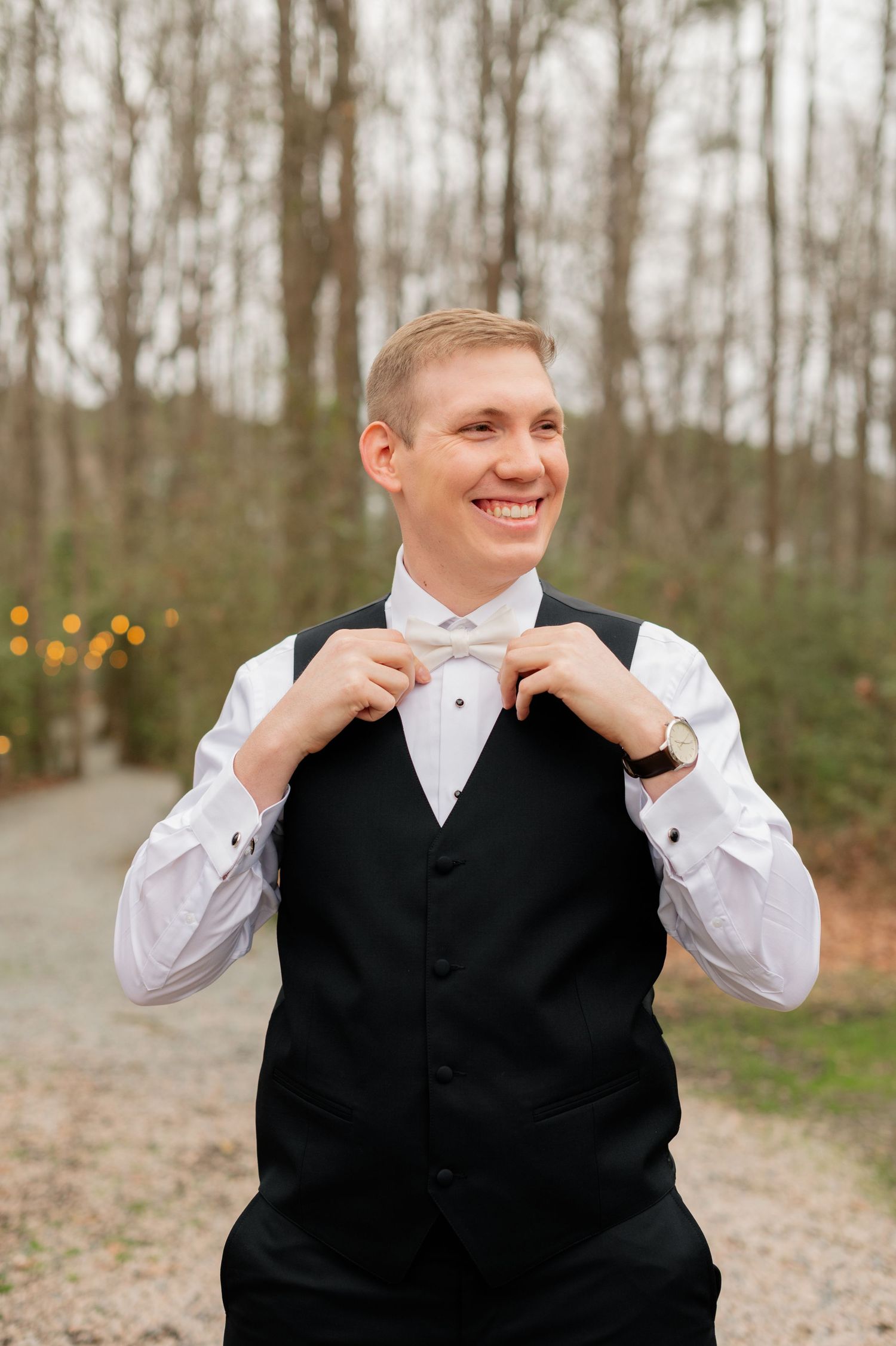 A groom adjusts his bowtie while smiling outdoors against a wooded background.