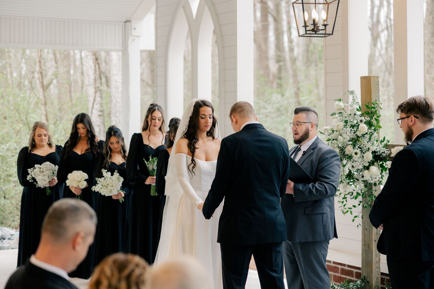 Elegant wedding ceremony in a bright white columned venue with guests watching and a floral-decorated altar area.