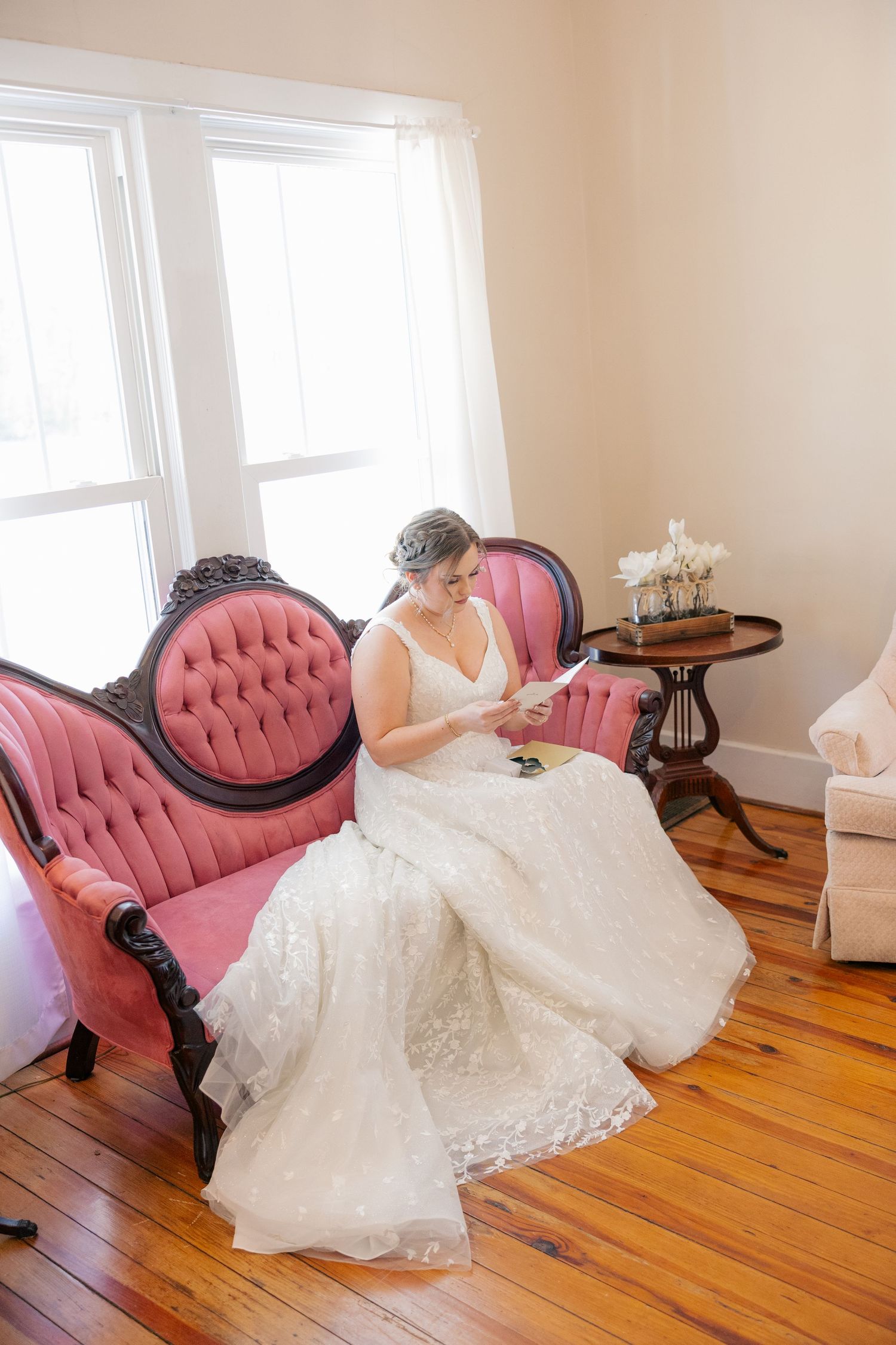 A white lace wedding dress flows over an elegant pink vintage chaise lounge chair on hardwood floors.