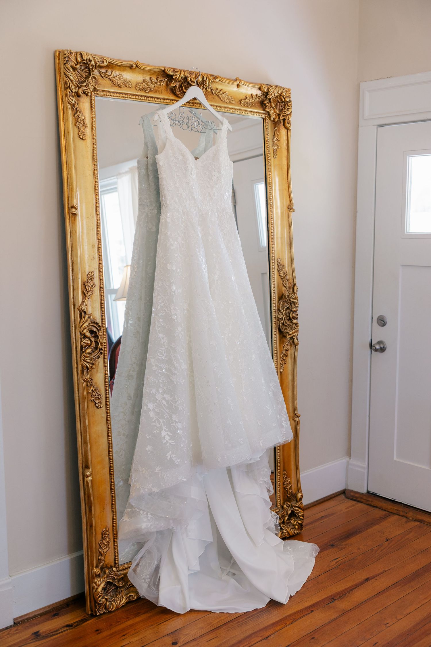 White lace wedding dress displayed on a hanger against an ornate gold mirror frame.