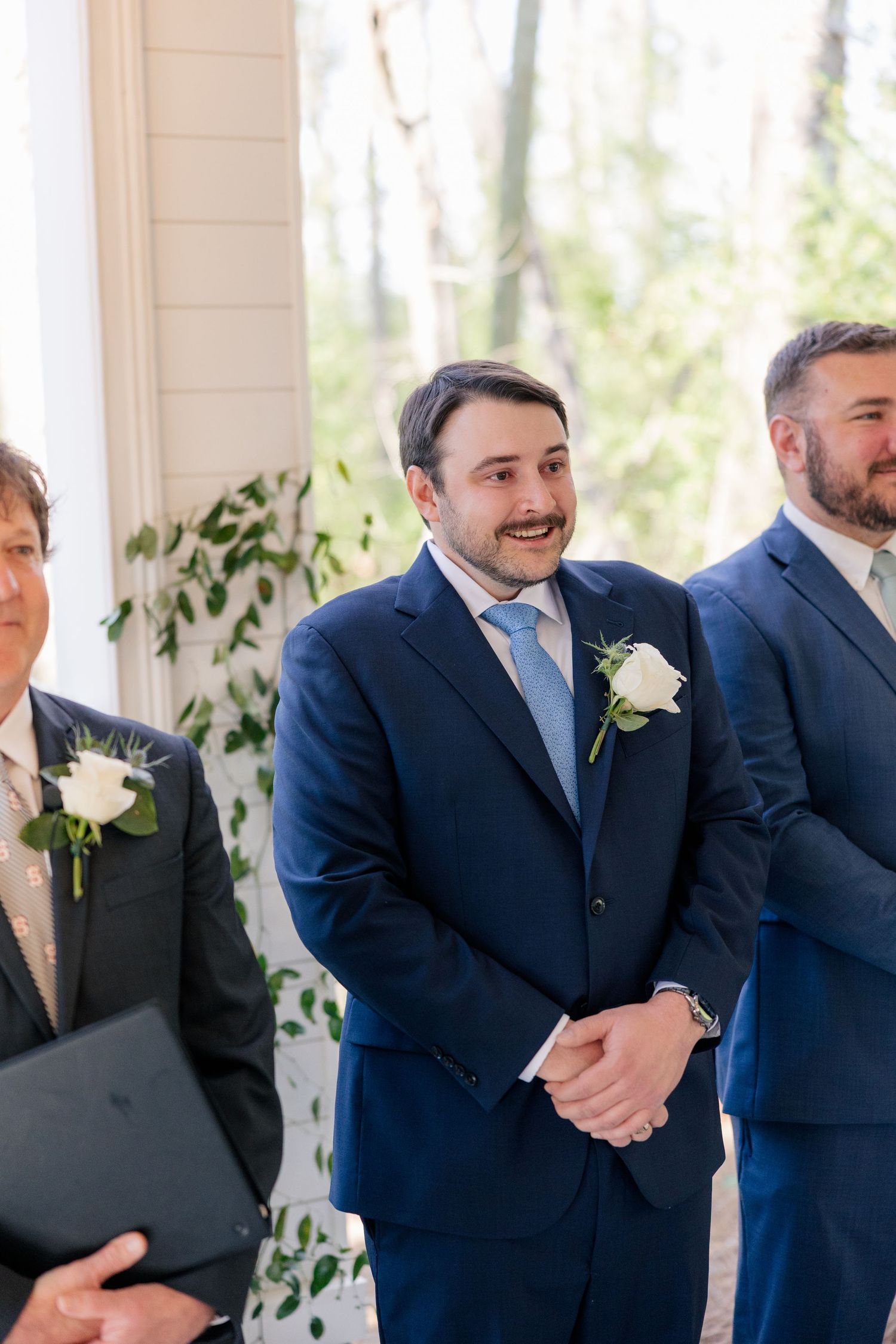 Groomsmen in navy blue suits with white boutonnieres smile during a wedding ceremony.