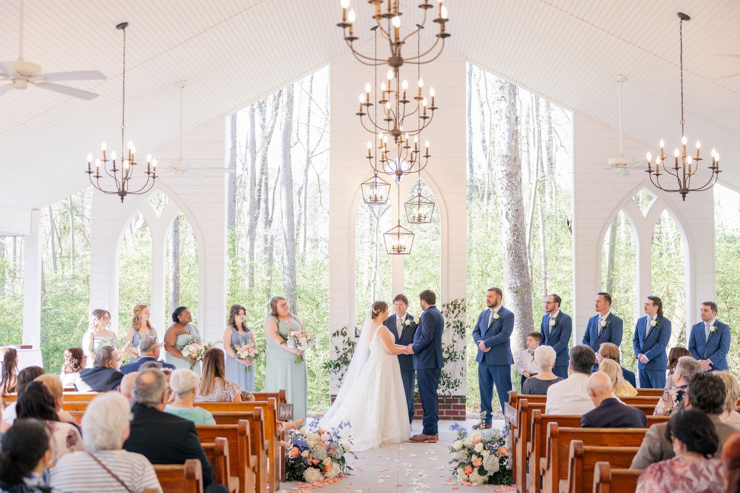 Wedding ceremony venue with wooden pews, chandeliers and large windows overlooking a wooded area.