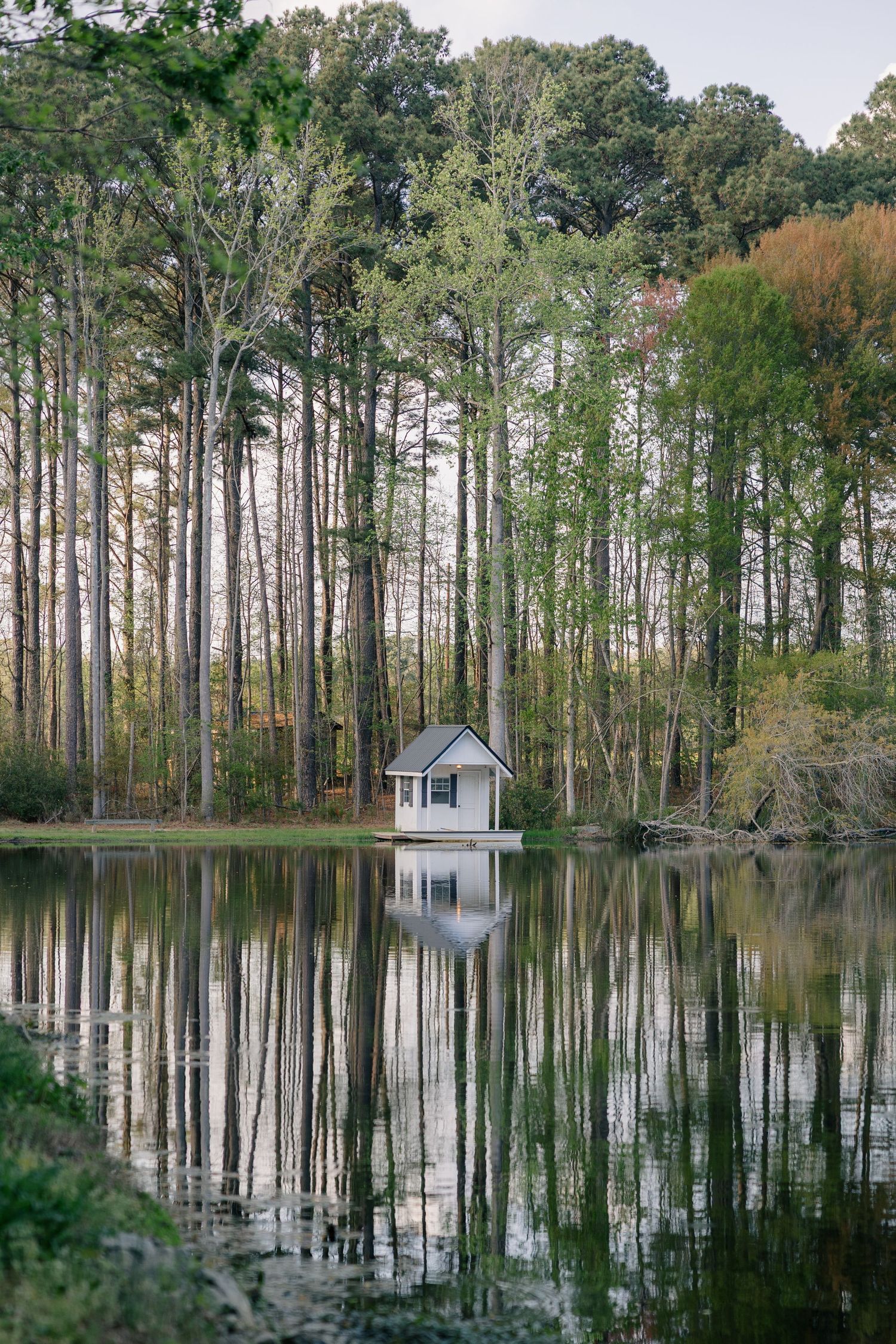 Small white boathouse reflected in still lake waters surrounded by tall pine trees.