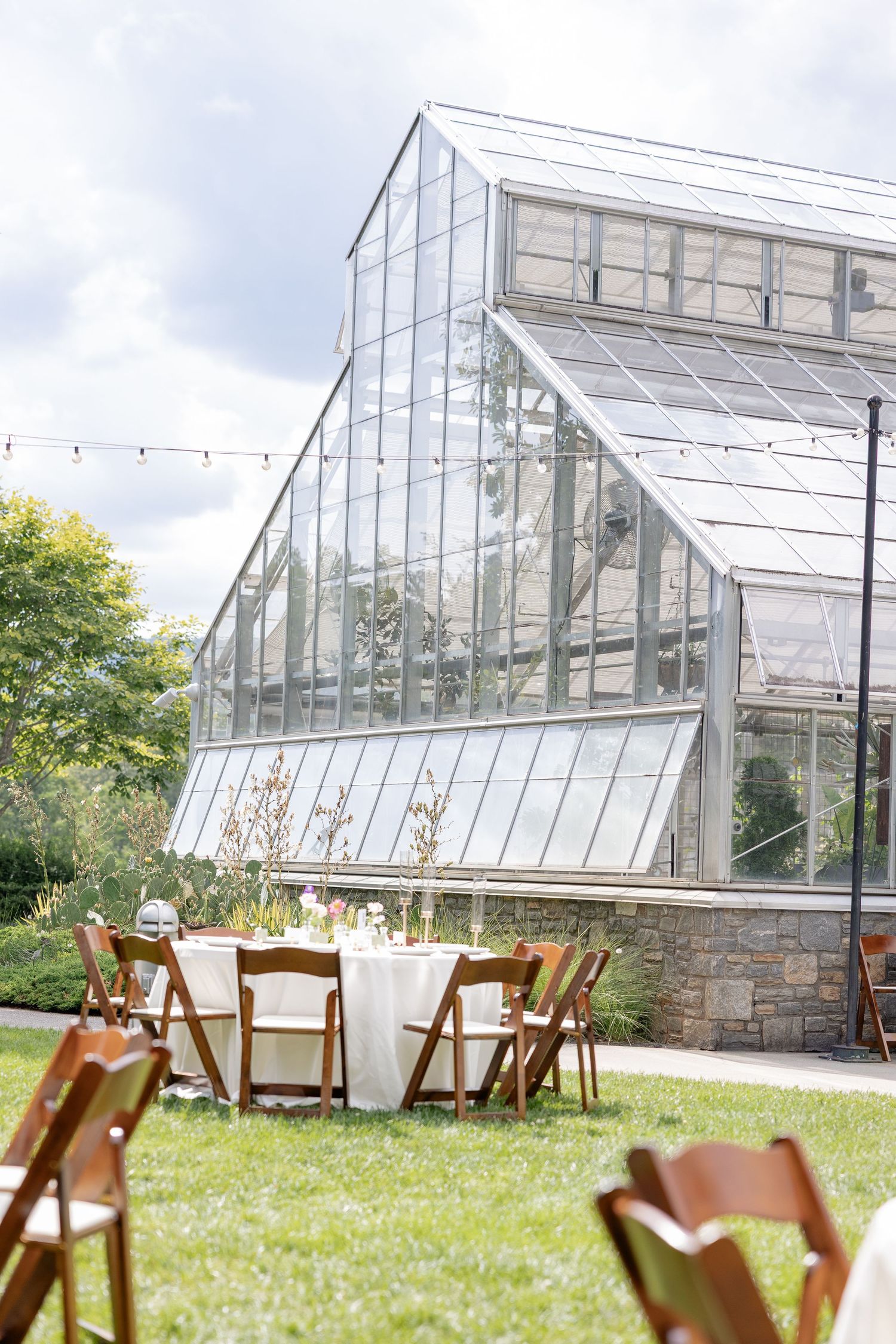 A large glass greenhouse structure sits in a grassy field with wooden chairs arranged outdoors.