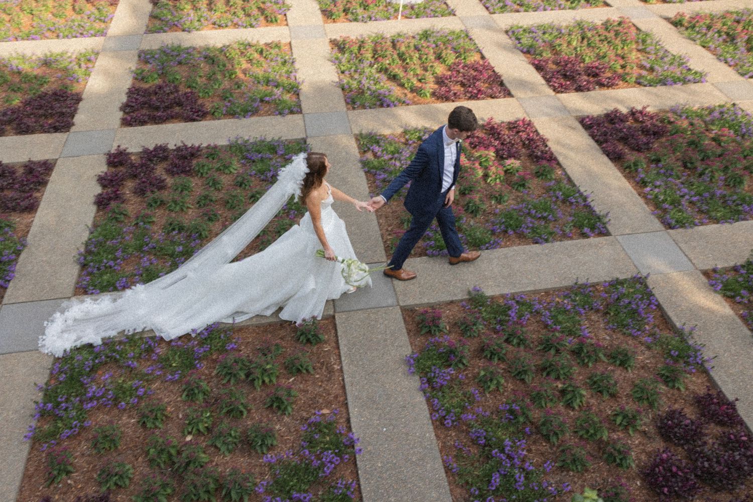 A couple in wedding attire walks hand in hand through a garden path with stone pavers and colorful landscaping.