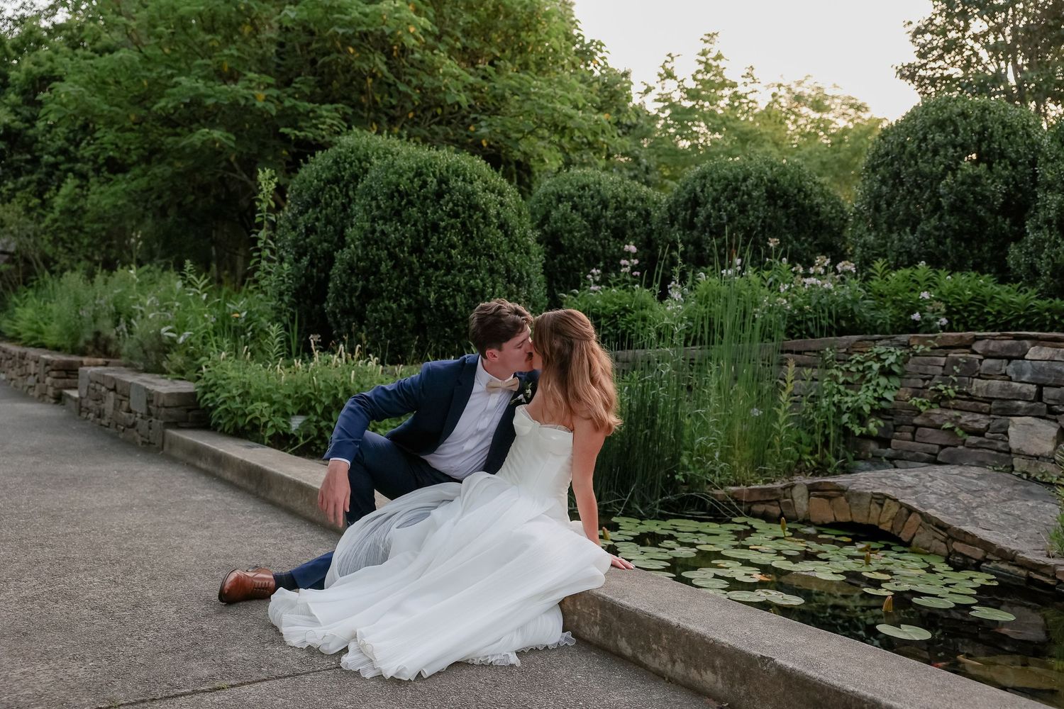 A romantic moment captured by a garden pond with stone walls and lush greenery.