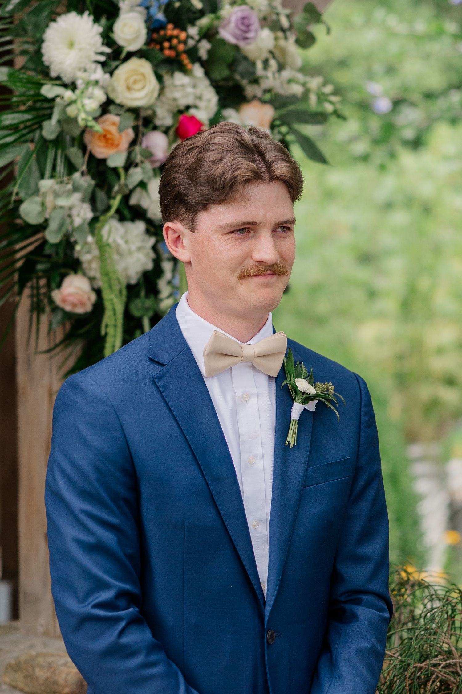 A groom in a navy suit and white tie stands in front of floral decorations at an outdoor wedding ceremony.
