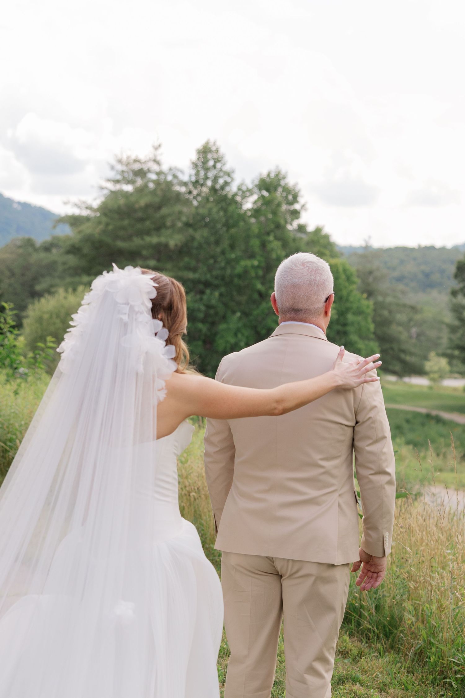 A bride in a flowing white dress and veil walks with someone in a beige suit through a garden setting.