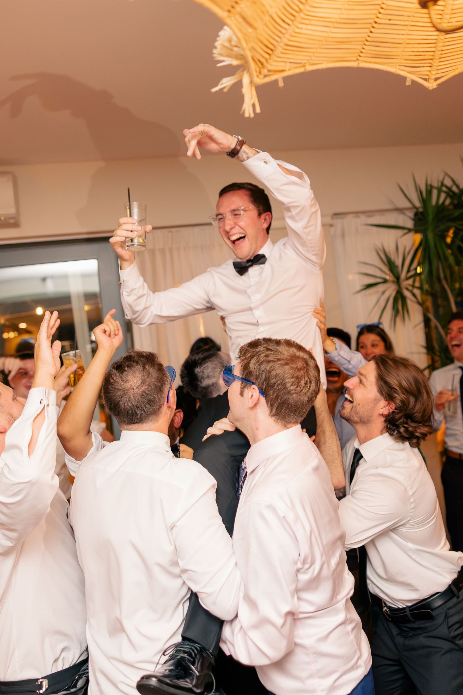 Excited groom lifted by groomsmen while holding champagne glass during lively wedding reception party.