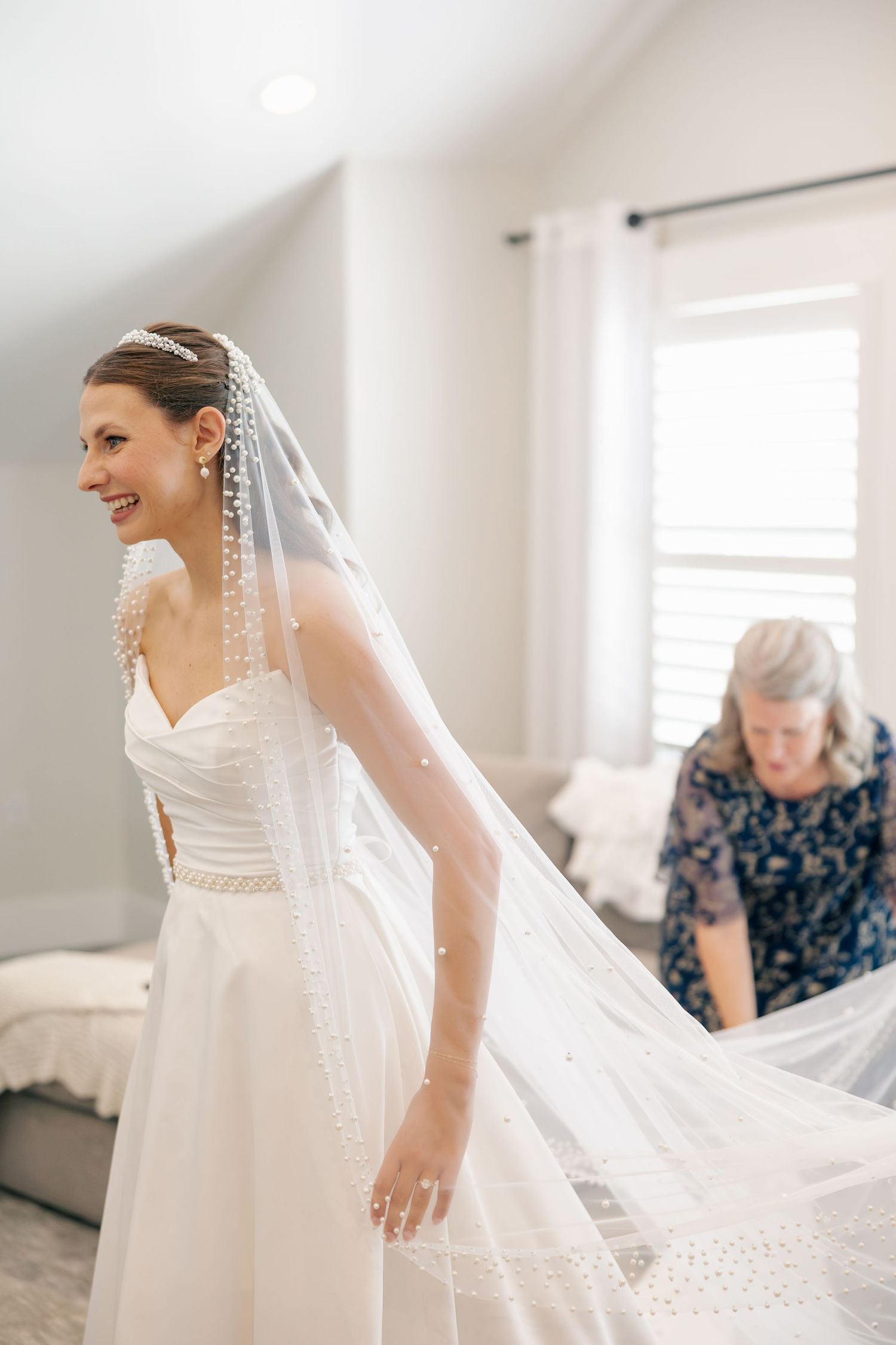 Bride smiles in strapless wedding gown with pearl-trimmed veil while mother adjusts train in bright room.