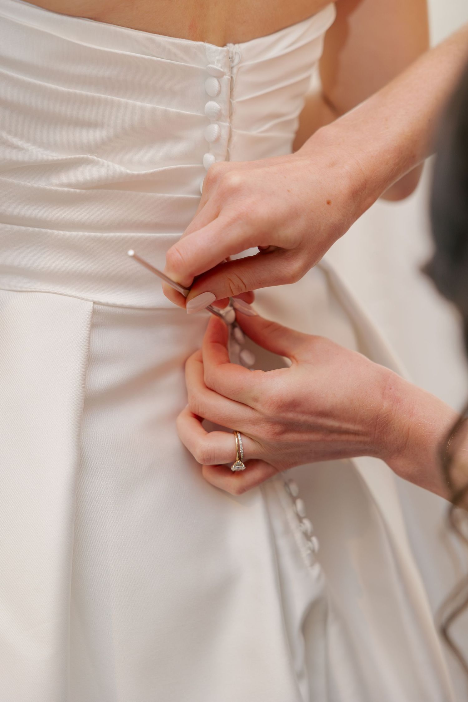 Hands carefully buttoning the back of a white satin wedding gown, with a diamond ring visible.