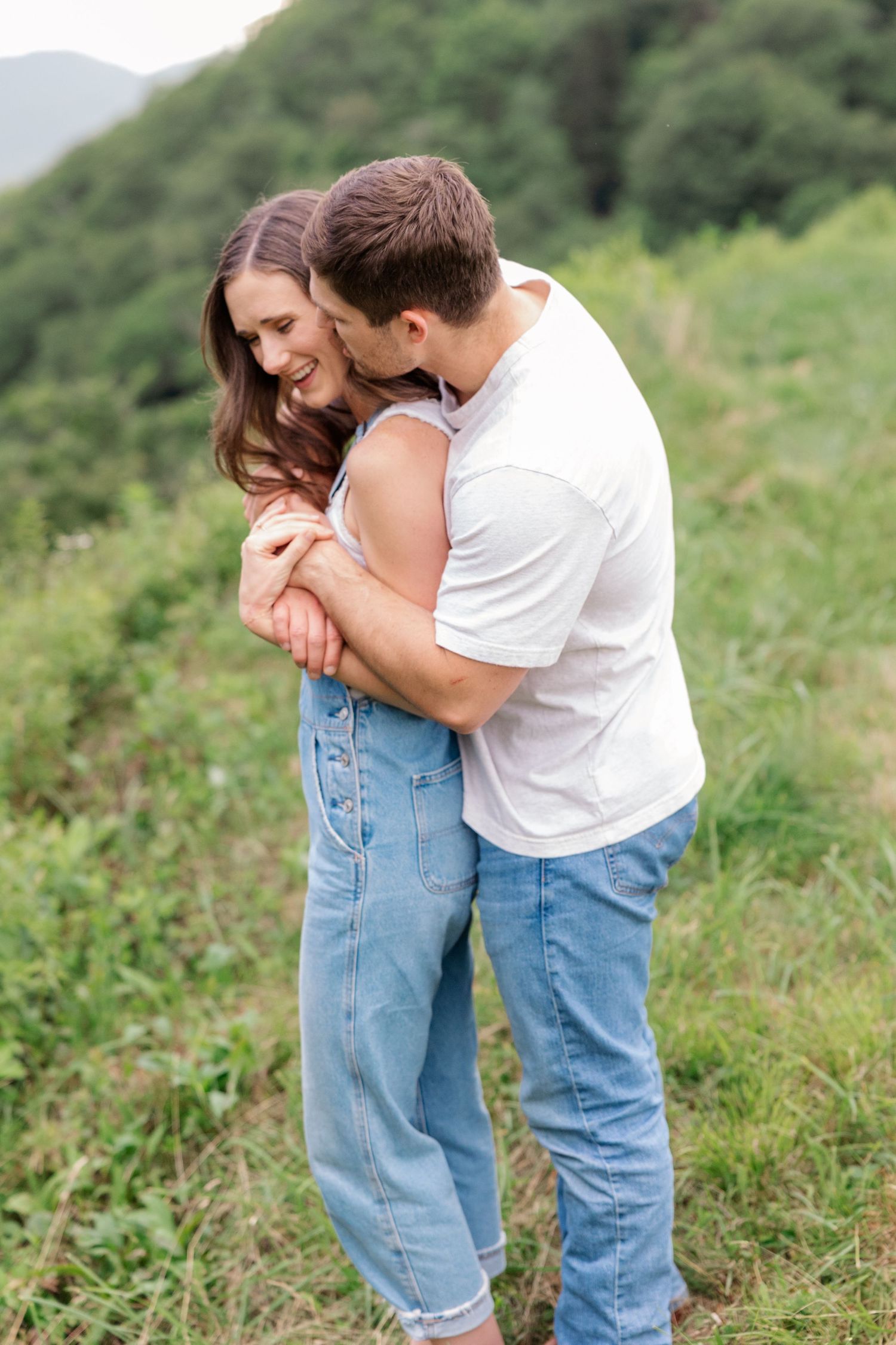 A romantic couple embracing in casual denim outfits against a lush green mountain backdrop.
