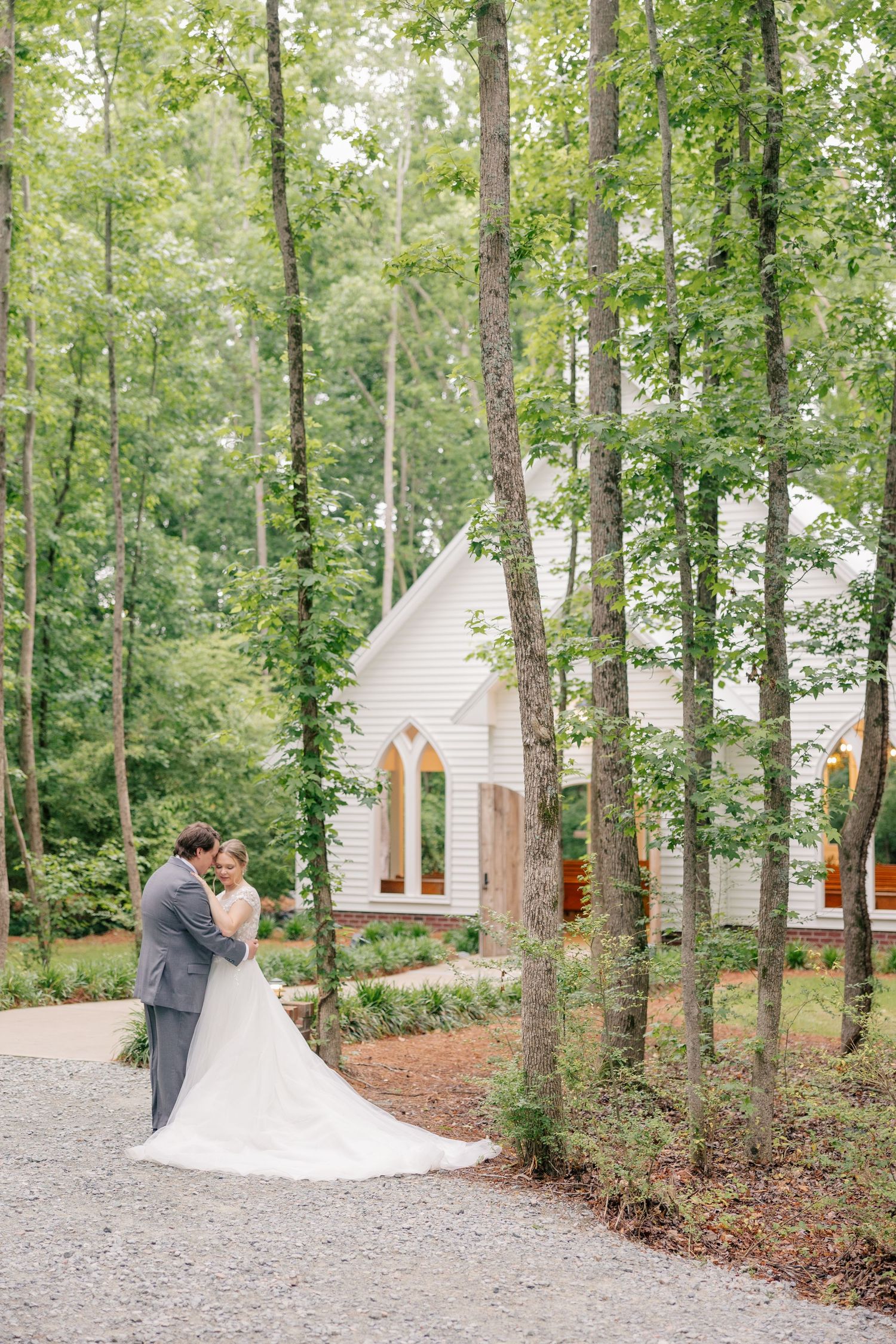 A bride and groom share a romantic embrace among tall trees beside a charming white chapel in a lush forest setting.