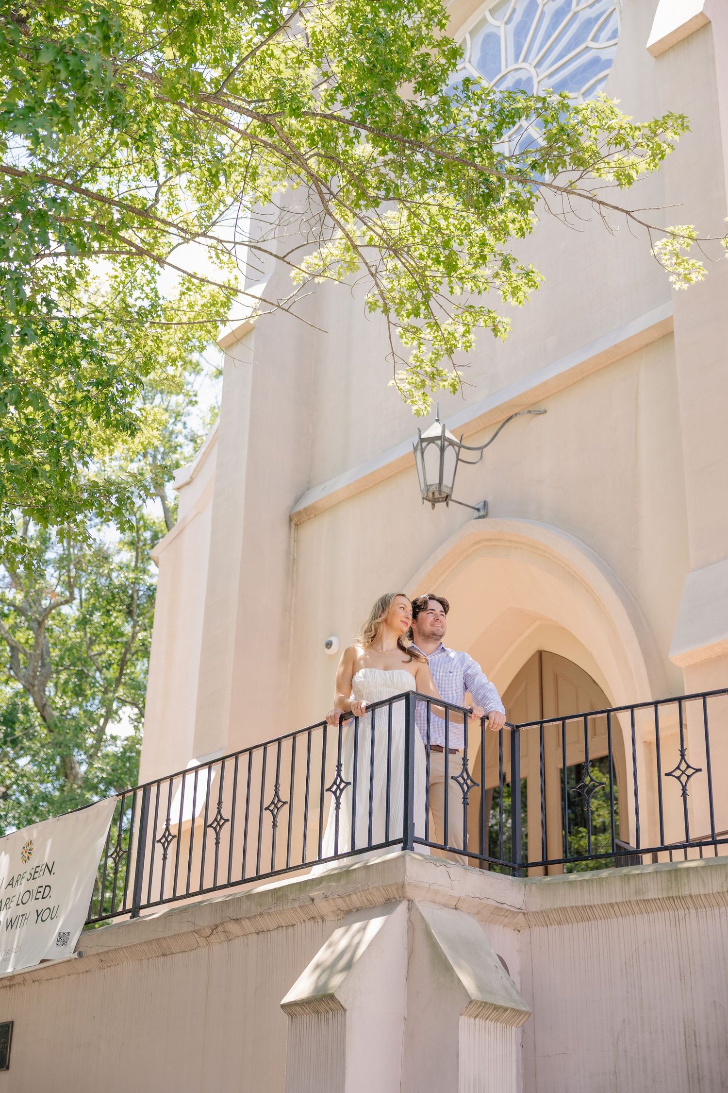 Couple embracing on a sunlit church balcony surrounded by lush green trees during an outdoor wedding photo session.