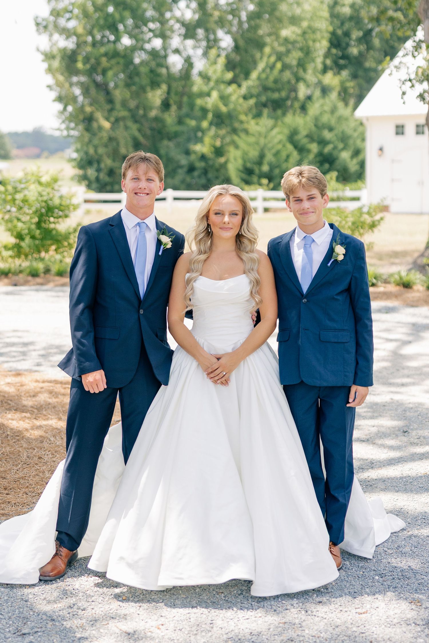 Bride in white gown poses with two groomsmen in navy suits outdoors at a wedding.