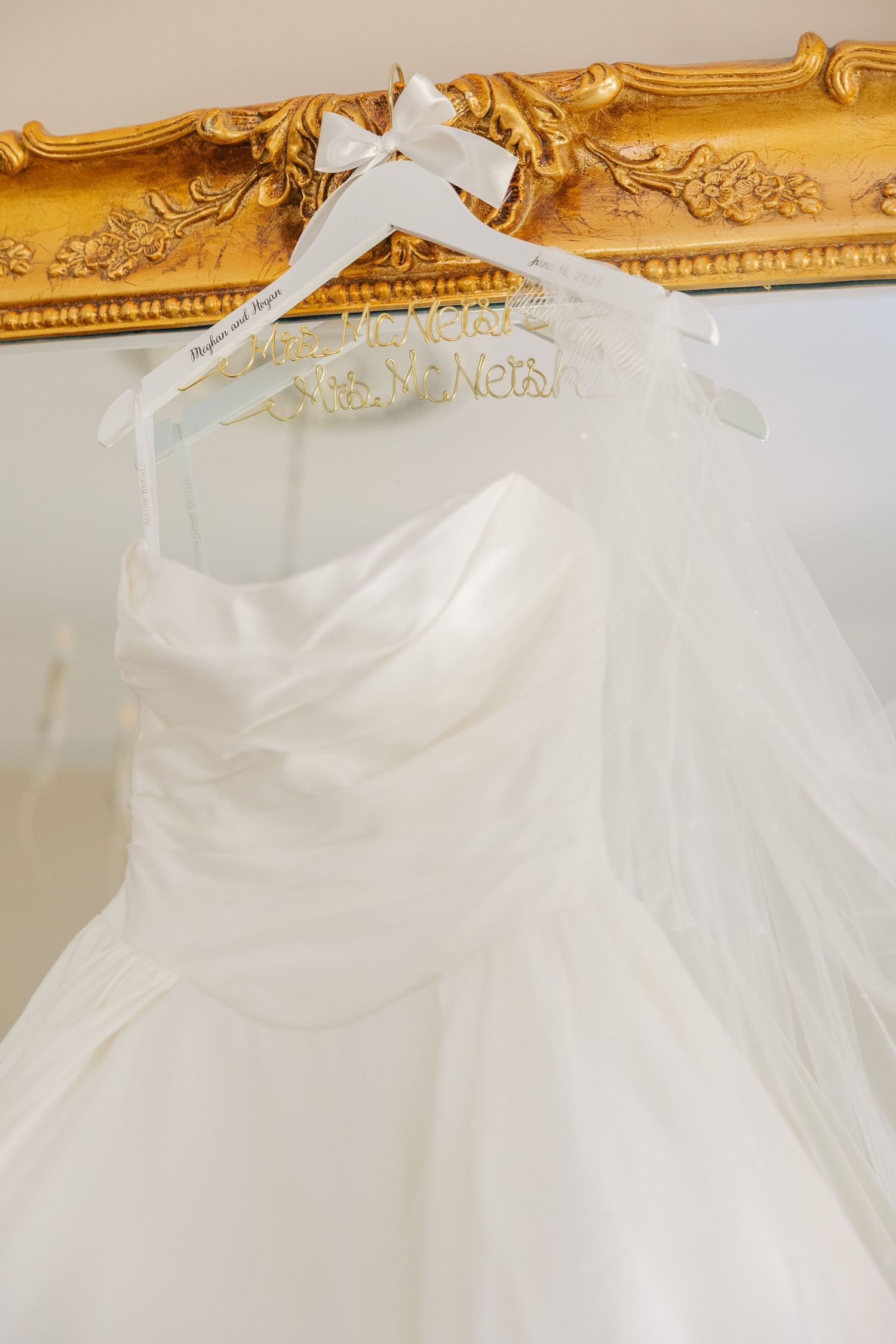 White wedding dress hanging on a decorative gold ornate mirror frame with a personalized bridal hanger.